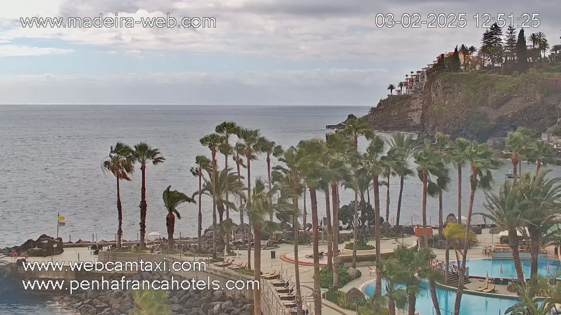 A resort with palm trees and swimming pools overlooks a windy ocean under a cloudy sky.