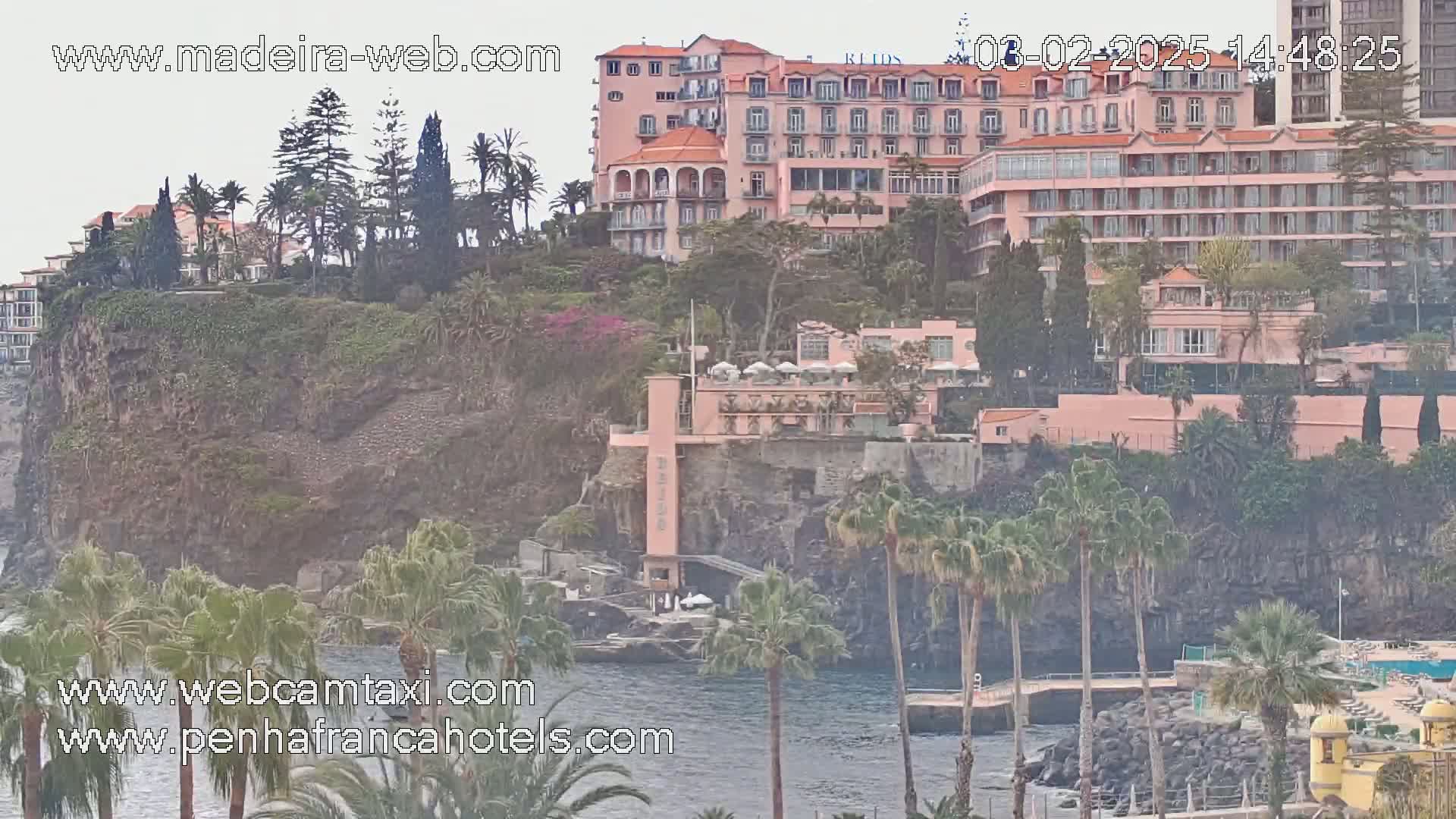 A pink hotel sits atop a dark cliff overlooking a calm body of water, surrounded by lush green vegetation and palm trees under an overcast sky.