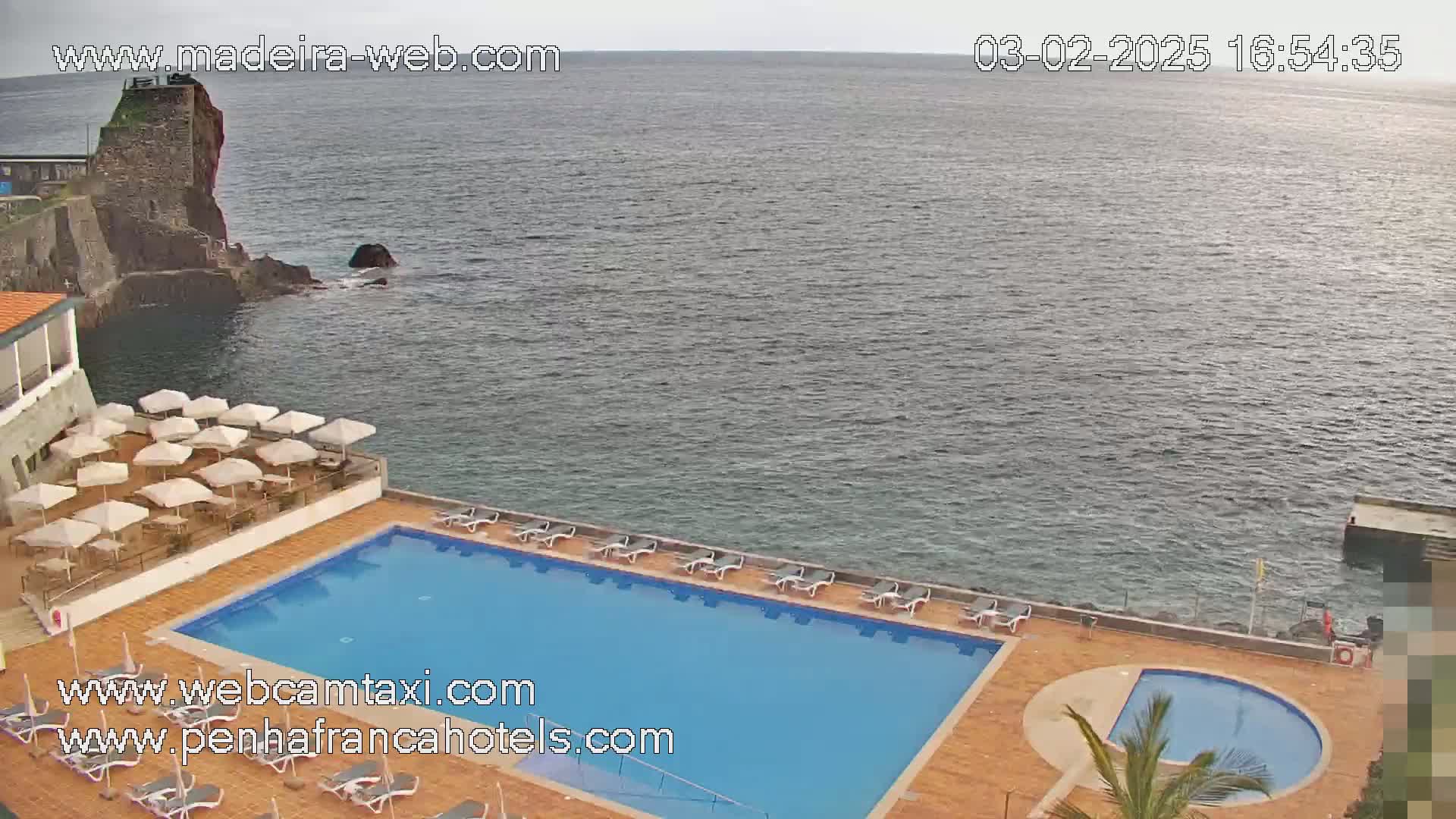 An outdoor swimming pool and patio area at a hotel overlook a calm ocean on an overcast day.