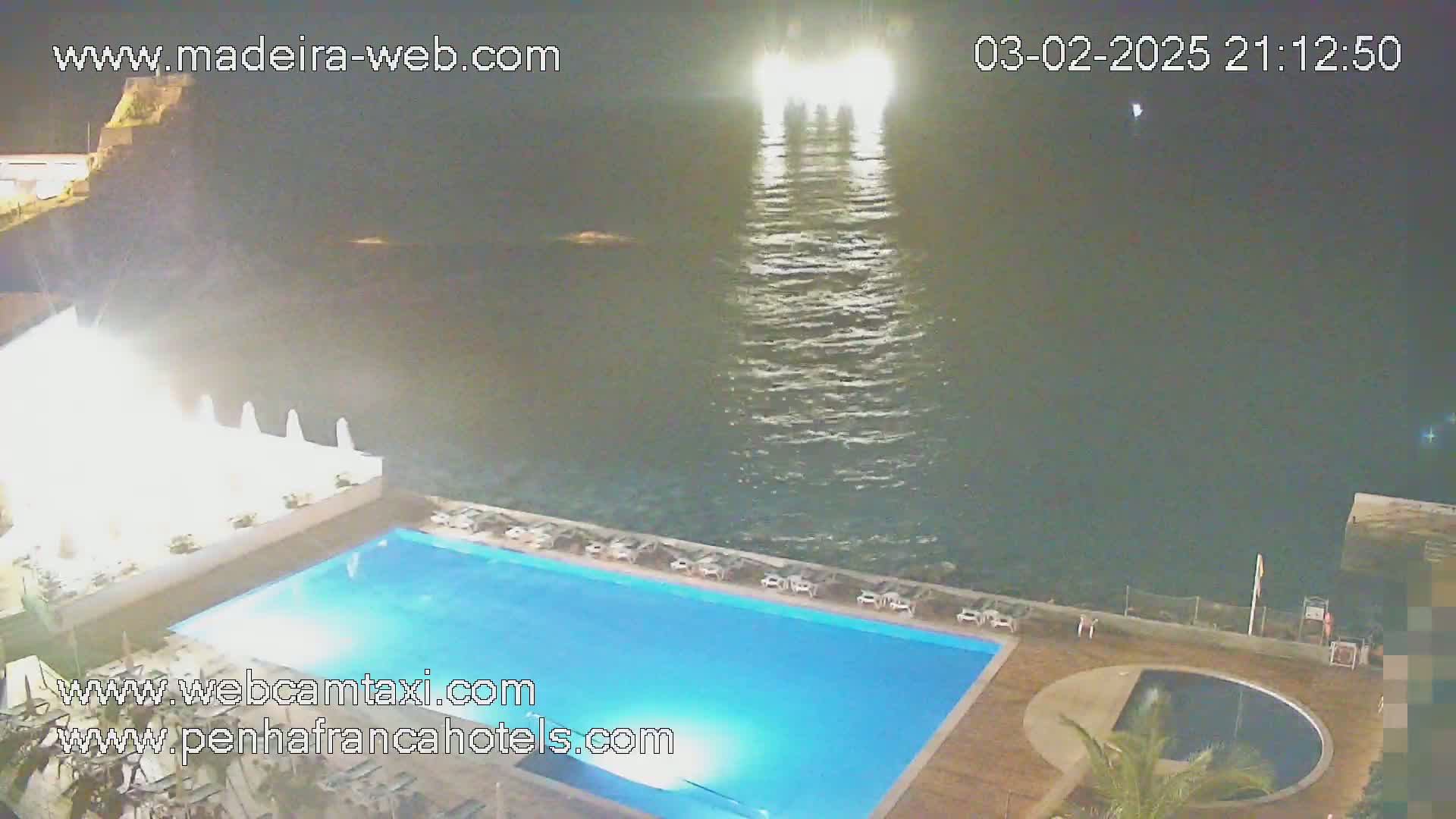 At night, a brightly lit outdoor swimming pool is adjacent to a dark ocean, with lights visible on the water.