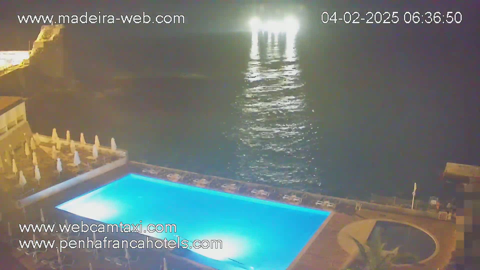 A nighttime view of a brightly lit swimming pool at a resort overlooking a dark ocean with faint lights reflecting on the water.