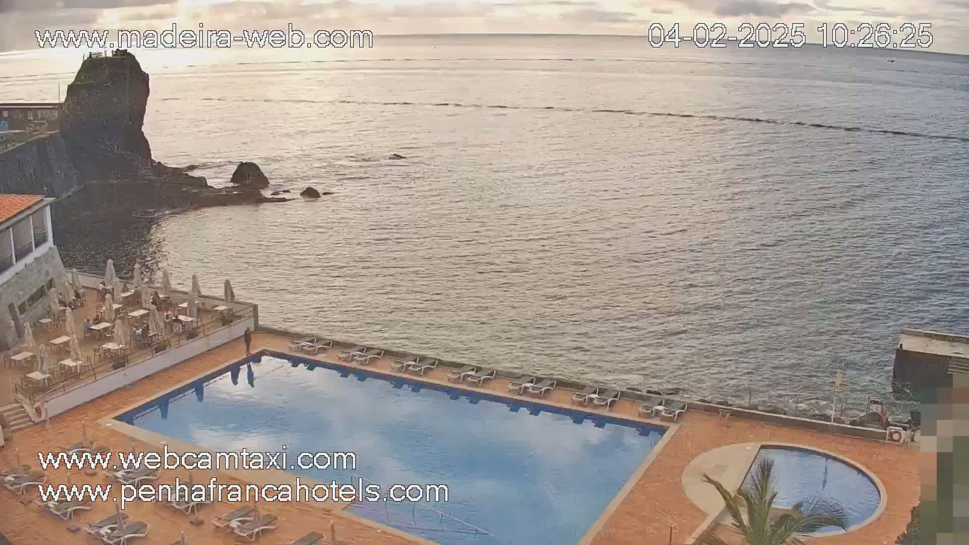 A hotel pool and patio area overlooking a calm ocean with a rocky shoreline under a partly cloudy sky.