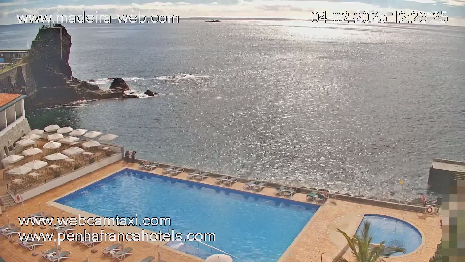 An outdoor swimming pool and a smaller adjacent pool are situated next to a rocky ocean shore on a partly sunny day.