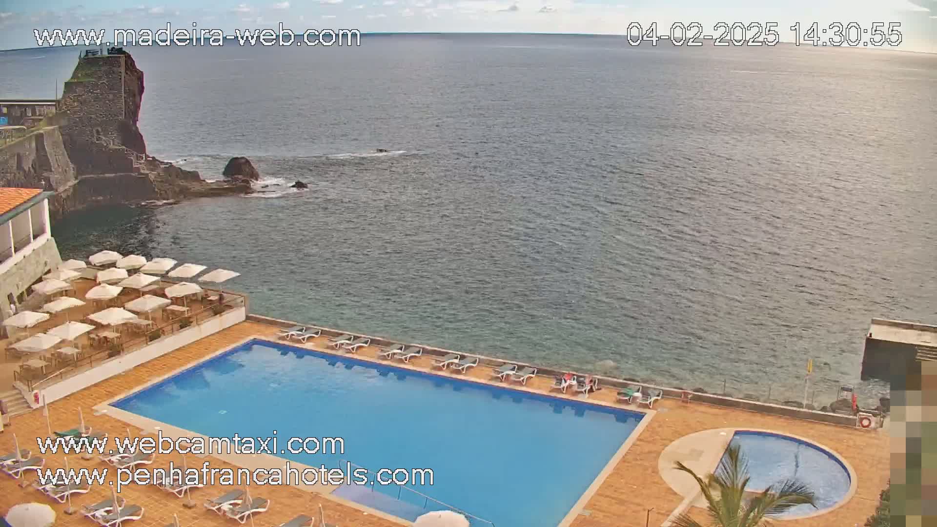 An outdoor swimming pool and lounge area are situated next to a rocky coast overlooking a calm ocean under a partly cloudy sky.