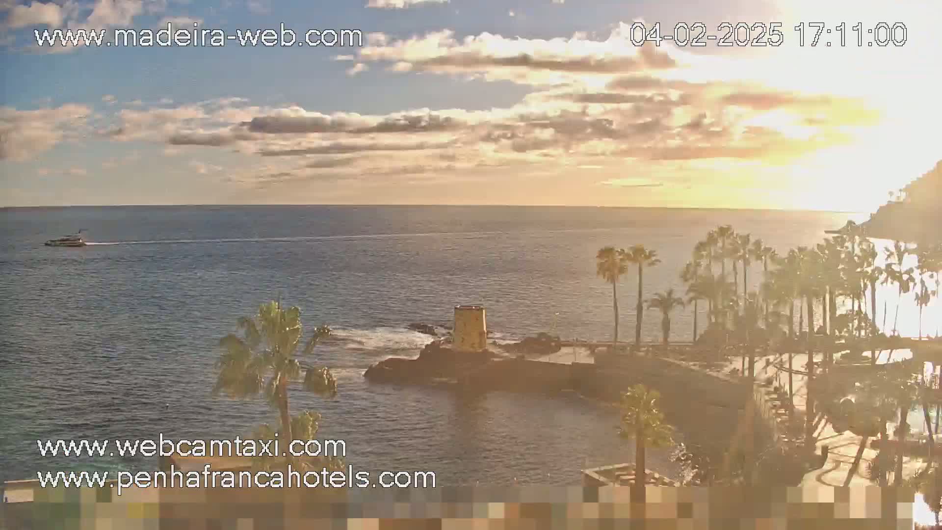 A calm ocean with a boat in the distance is seen at sunset under a partly cloudy sky, with palm trees and a small tower on a rocky outcrop in the foreground.
