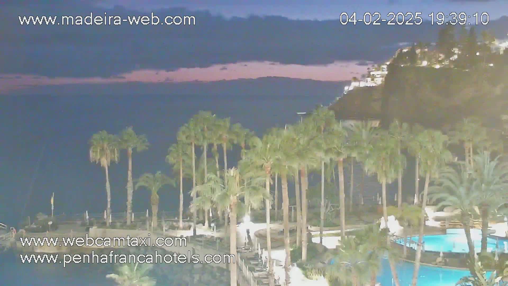 An ocean view at twilight shows palm trees surrounding a resort pool area situated on a cliff overlooking the water.