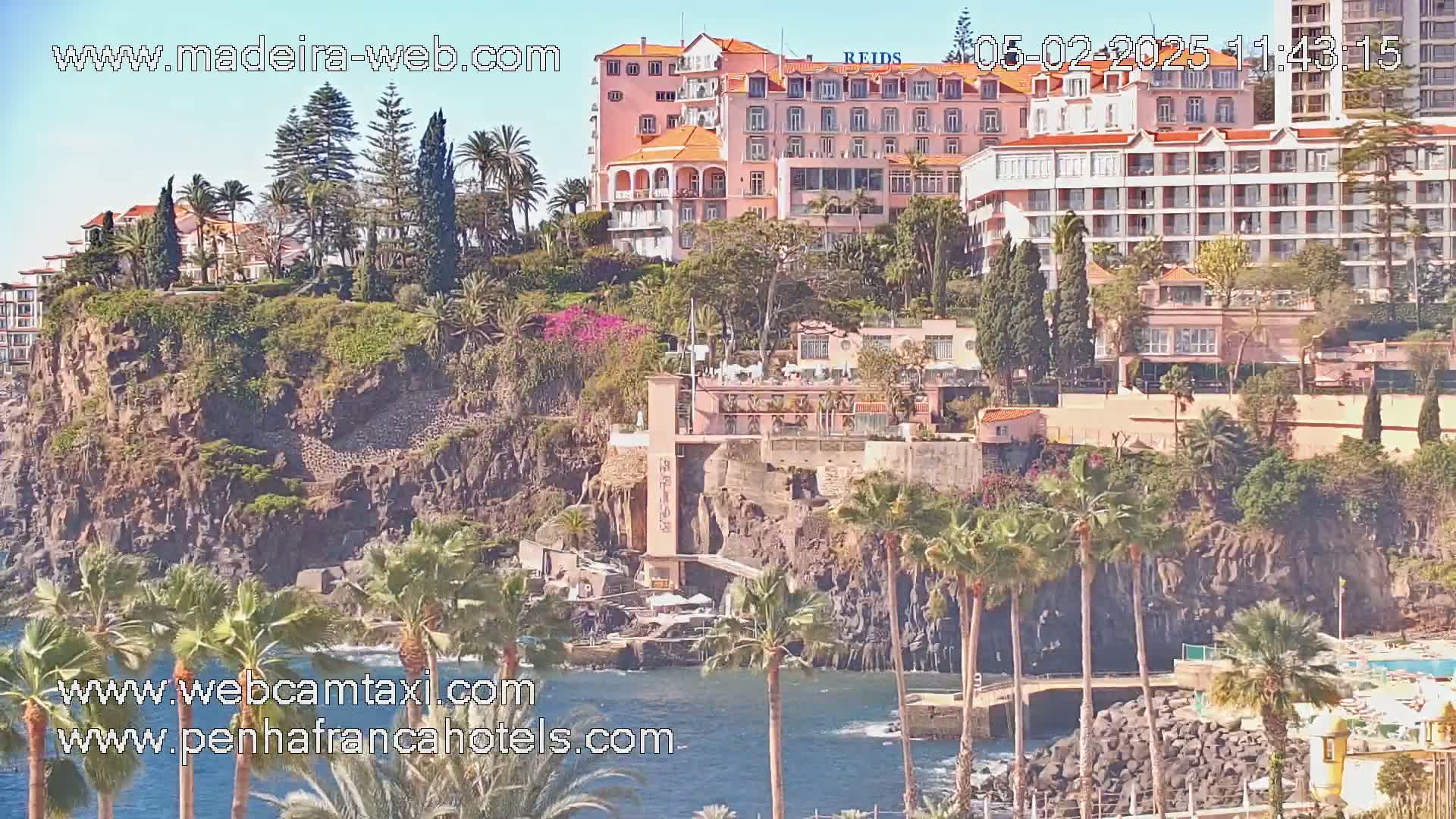 A rocky cliffside, dotted with palm trees and buildings, overlooks a calm body of water on a sunny day.