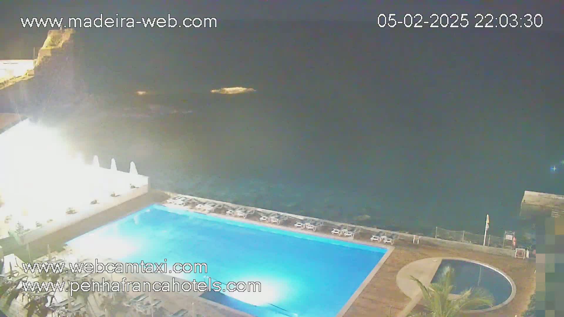 An illuminated swimming pool and a smaller circular pool are situated next to a dark ocean at night, with white lounge chairs surrounding the main pool.