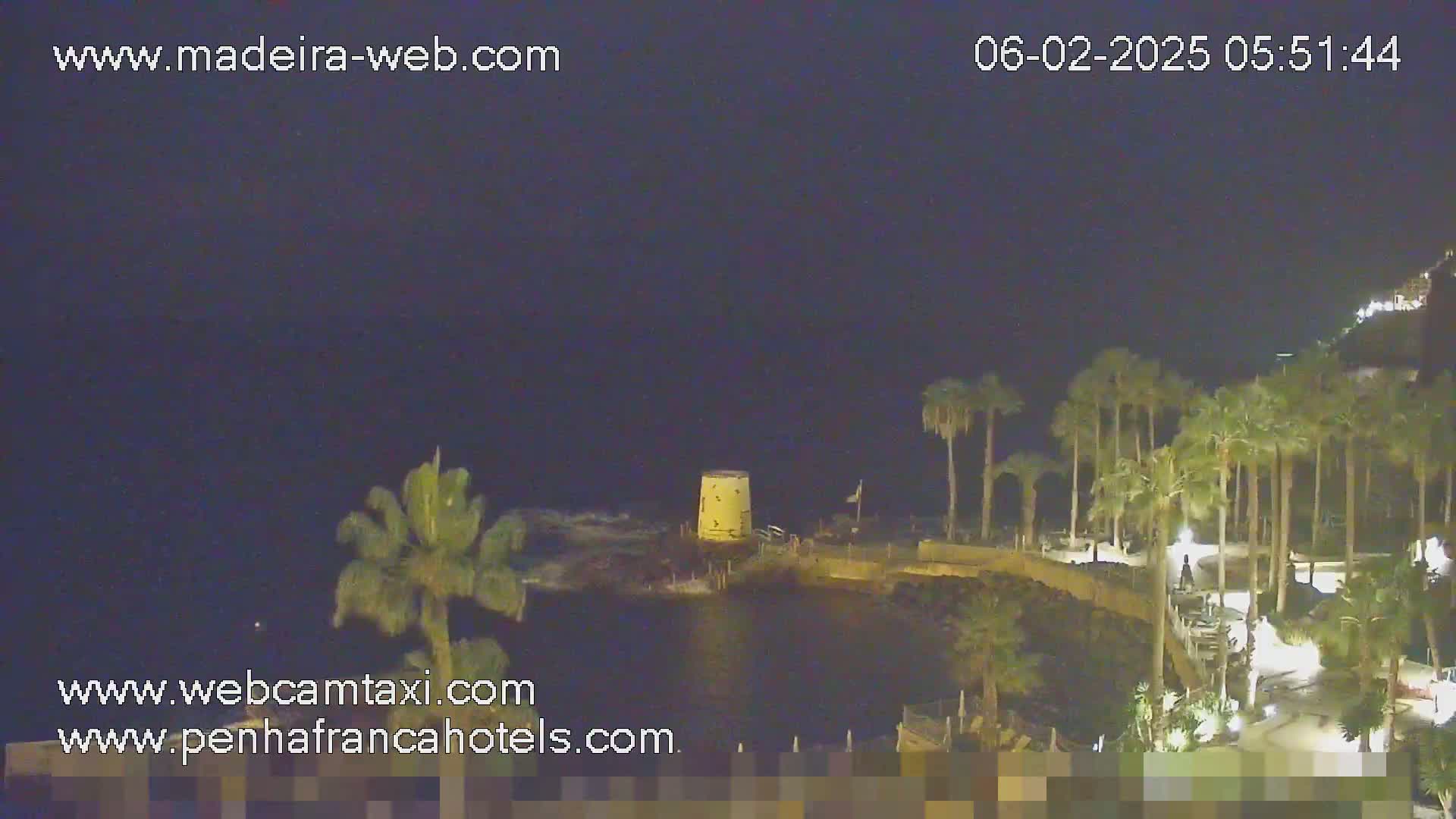 A nighttime coastal scene shows a rocky shoreline with palm trees, a small structure, and a dark ocean under a clear, dark sky.