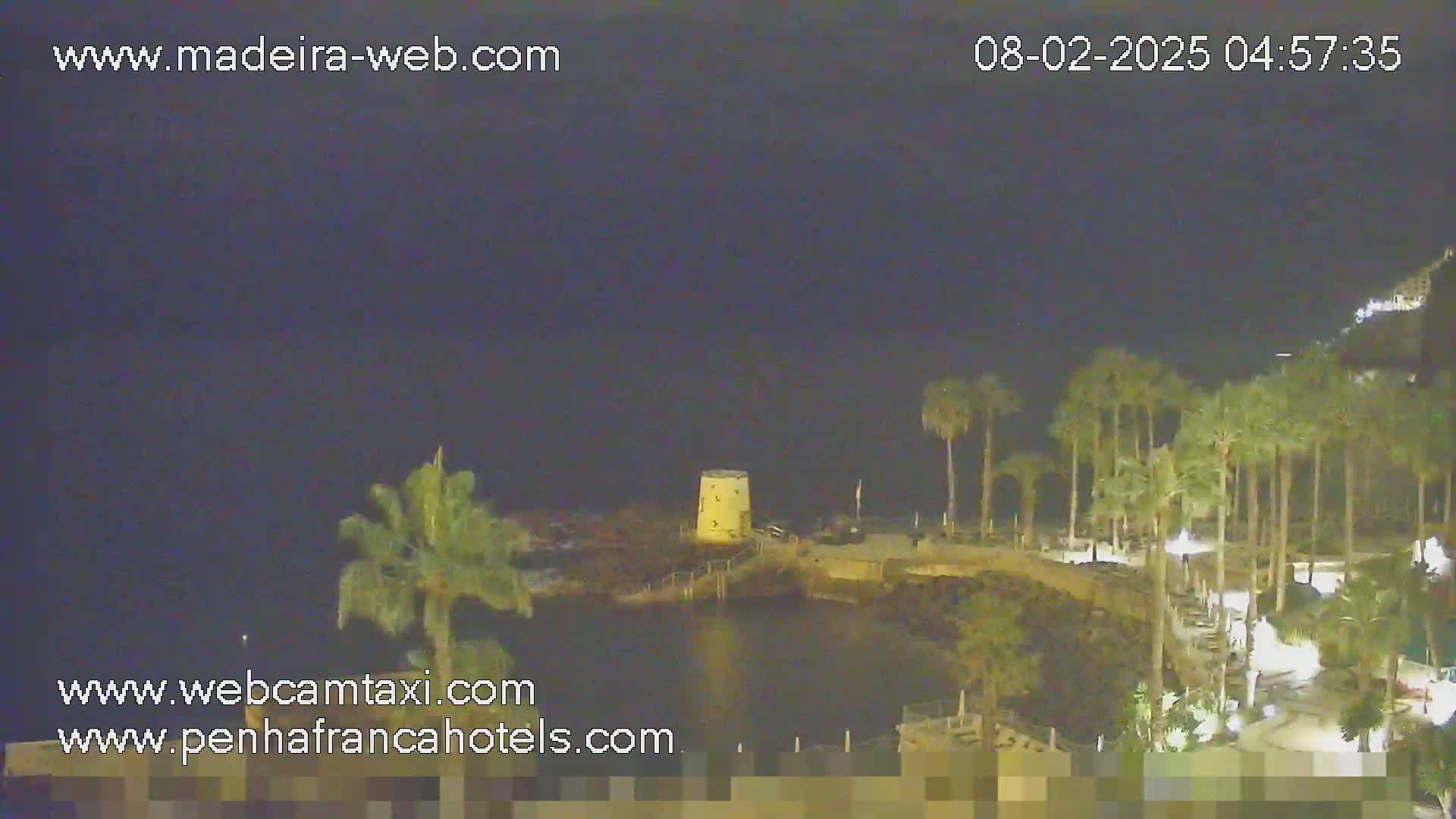 A nighttime, seaside view shows a small, illuminated tower on a rocky outcropping surrounded by palm trees and a walkway, under a dark sky.