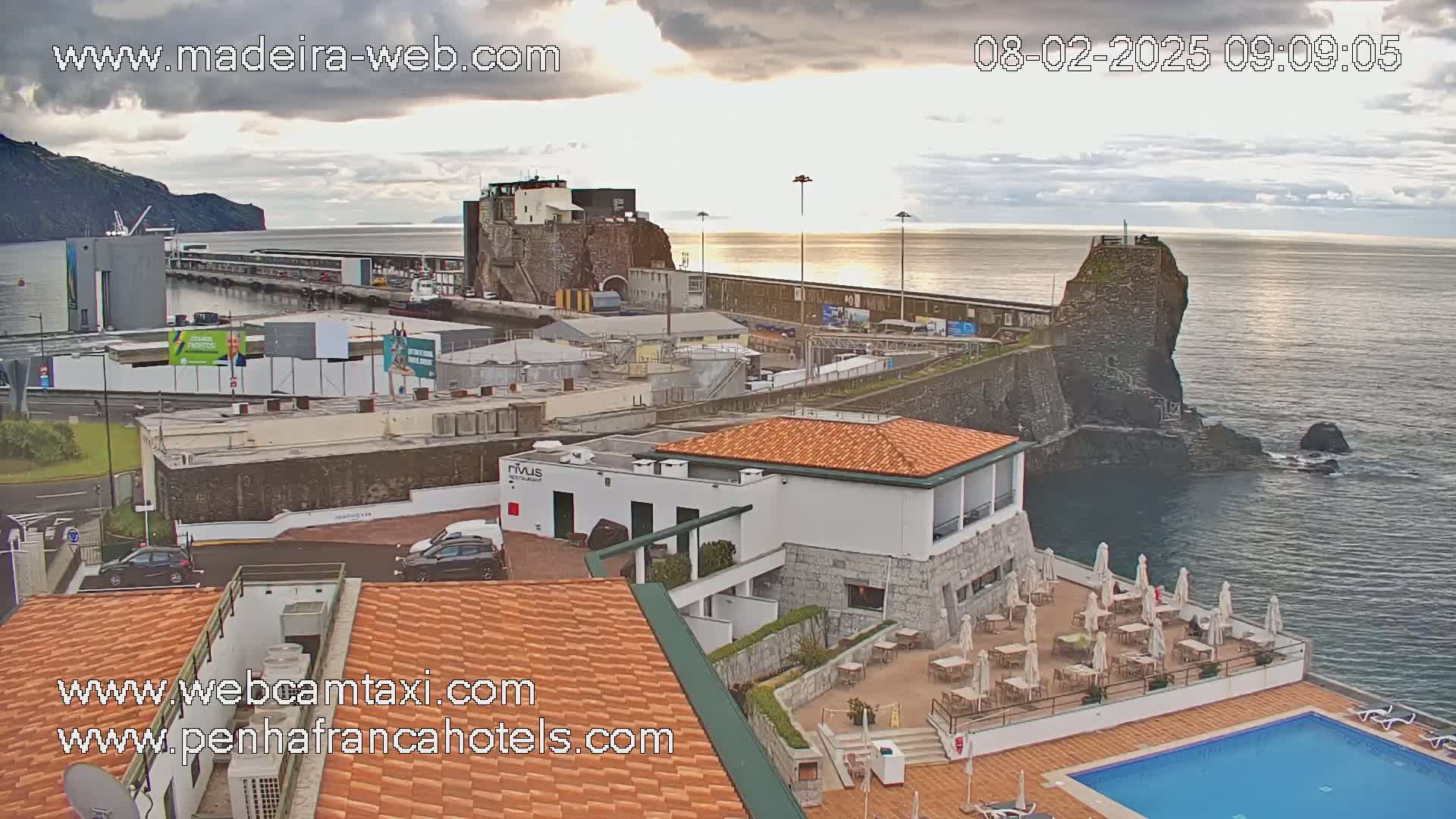 An overcast day shows a coastal town with a harbor, stone buildings, and a hotel with an outdoor pool overlooking a calm ocean.