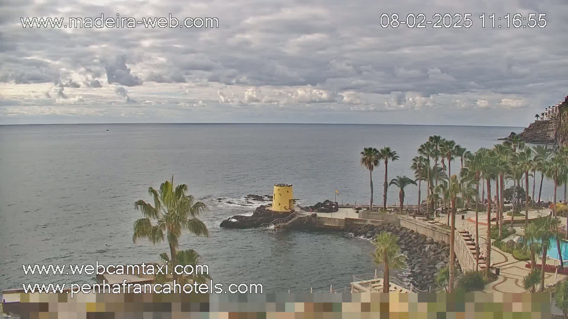 A partly cloudy day shows a calm ocean with a rocky shoreline lined with palm trees and a small yellow structure on a rock outcropping.