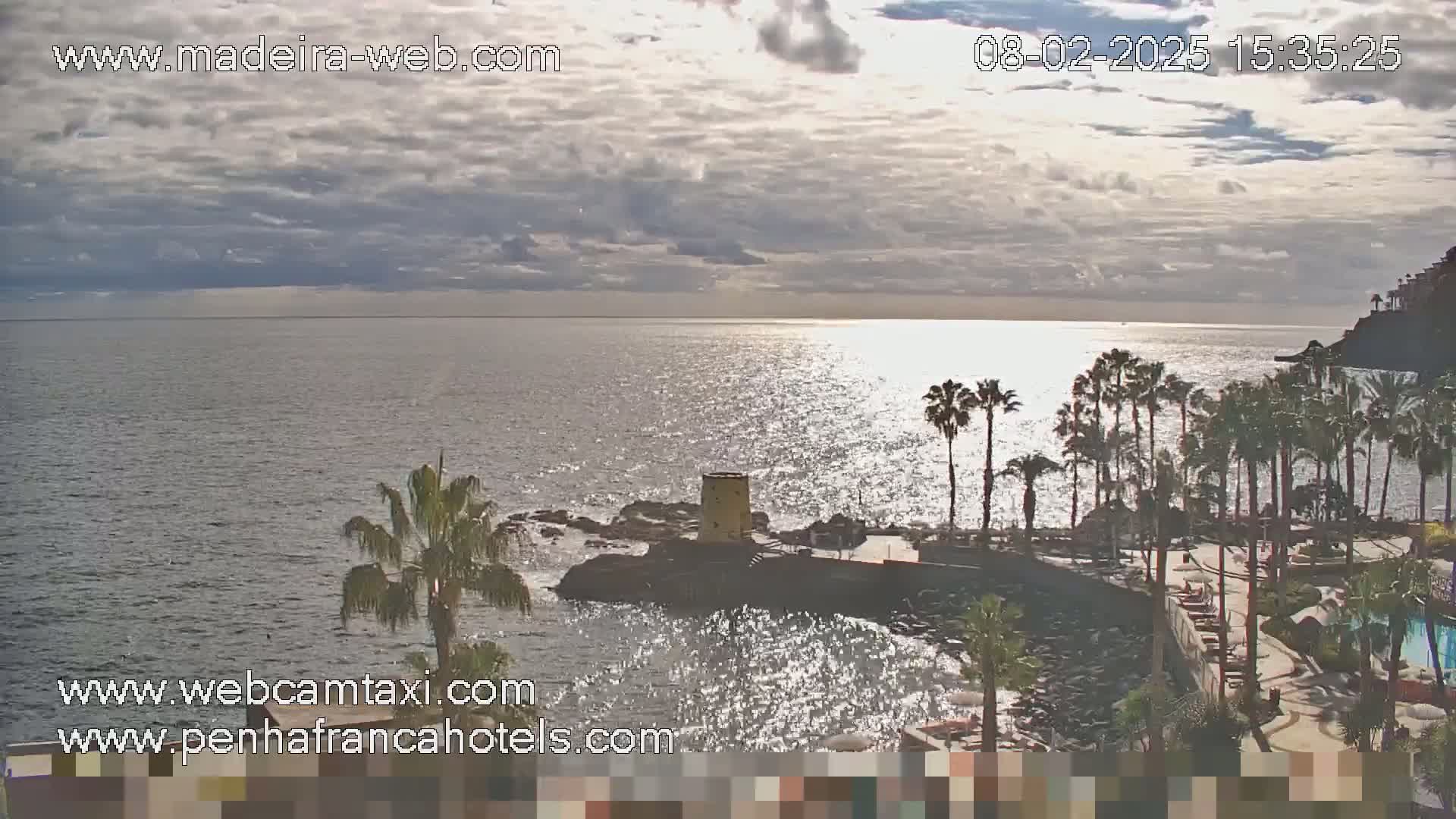 A partly cloudy day overlooks a calm ocean with a rocky shoreline dotted with palm trees and a small building.
