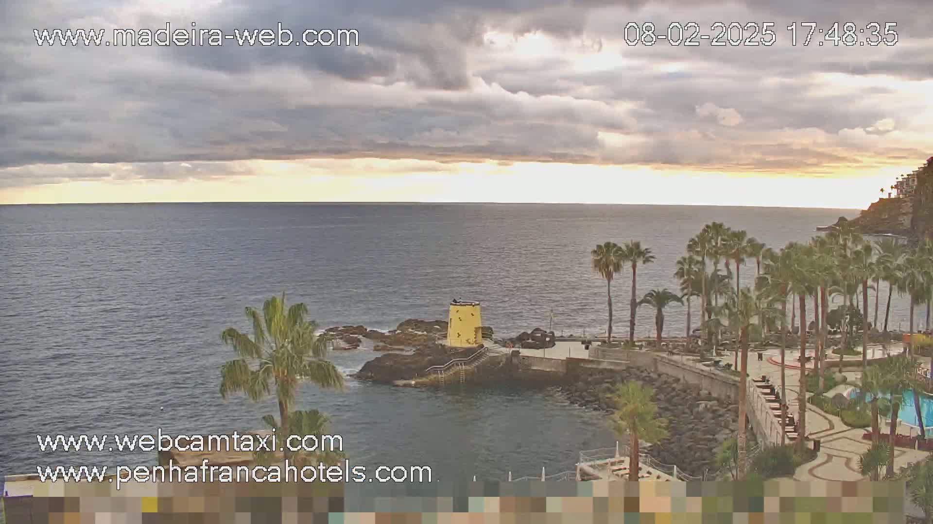 A partly cloudy sunset over a calm ocean, viewed from a rocky coastline with palm trees and resort buildings.