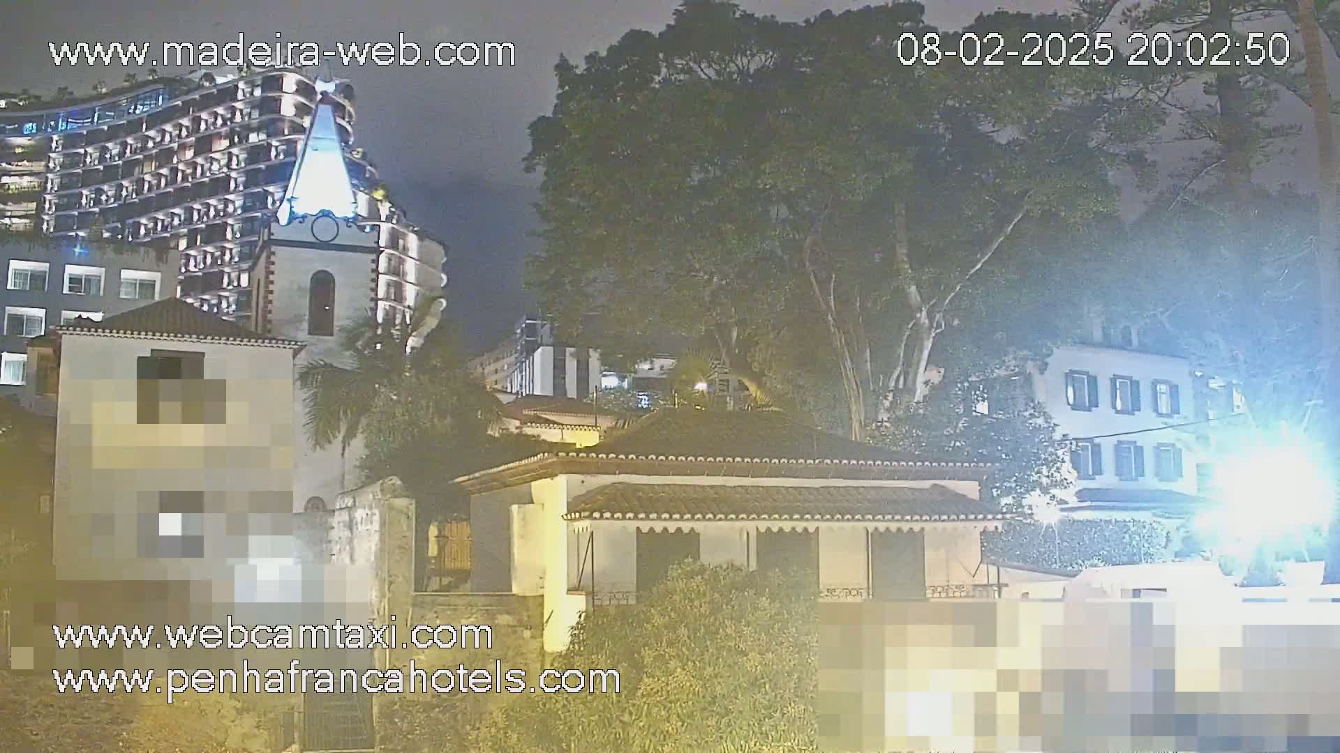 A nighttime view of buildings, including a tall building, a church-like structure, and smaller houses, under a partly cloudy sky with some trees.