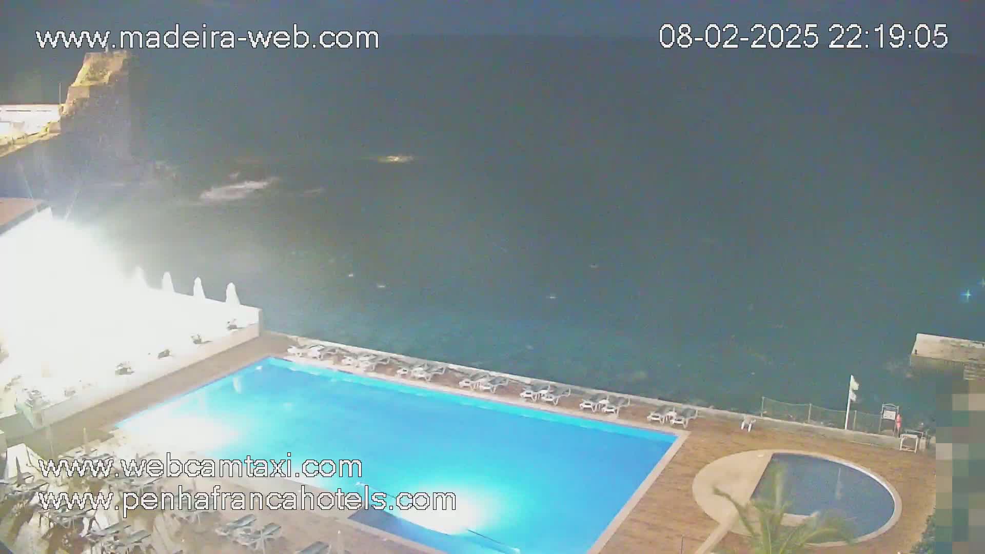 A nighttime view of a rectangular swimming pool and a smaller round pool next to it, overlooking a dark ocean.