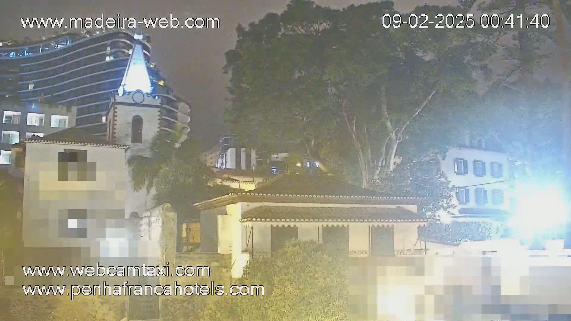 A nighttime view of buildings, including a tall modern structure and smaller, older buildings, partially obscured by trees under a dark, hazy sky.