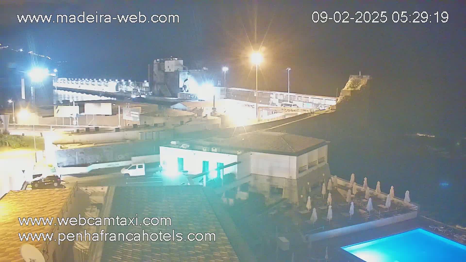 A nighttime view of a coastal town with a swimming pool and several buildings illuminated by artificial light, under a clear, dark sky.