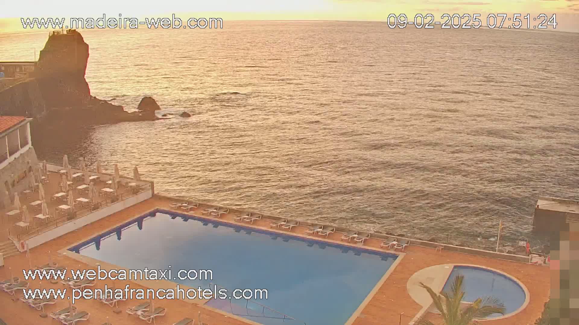 A hotel pool and patio area overlooks a calm ocean at sunrise.