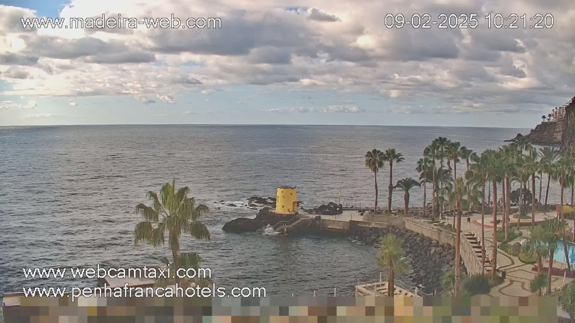 A partly cloudy day shows a calm ocean scene with palm trees lining a rocky shoreline and a small yellow structure near the water.