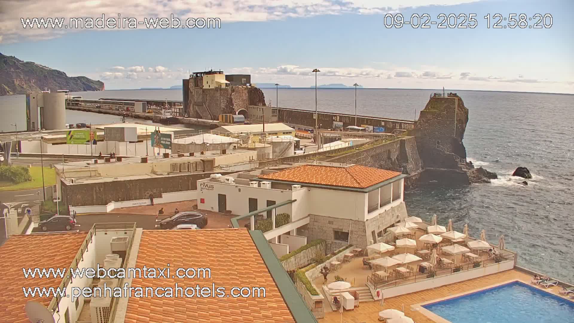 A partly cloudy day overlooks a harbor town with a rocky coastline, featuring buildings, a swimming pool, and outdoor patio seating.