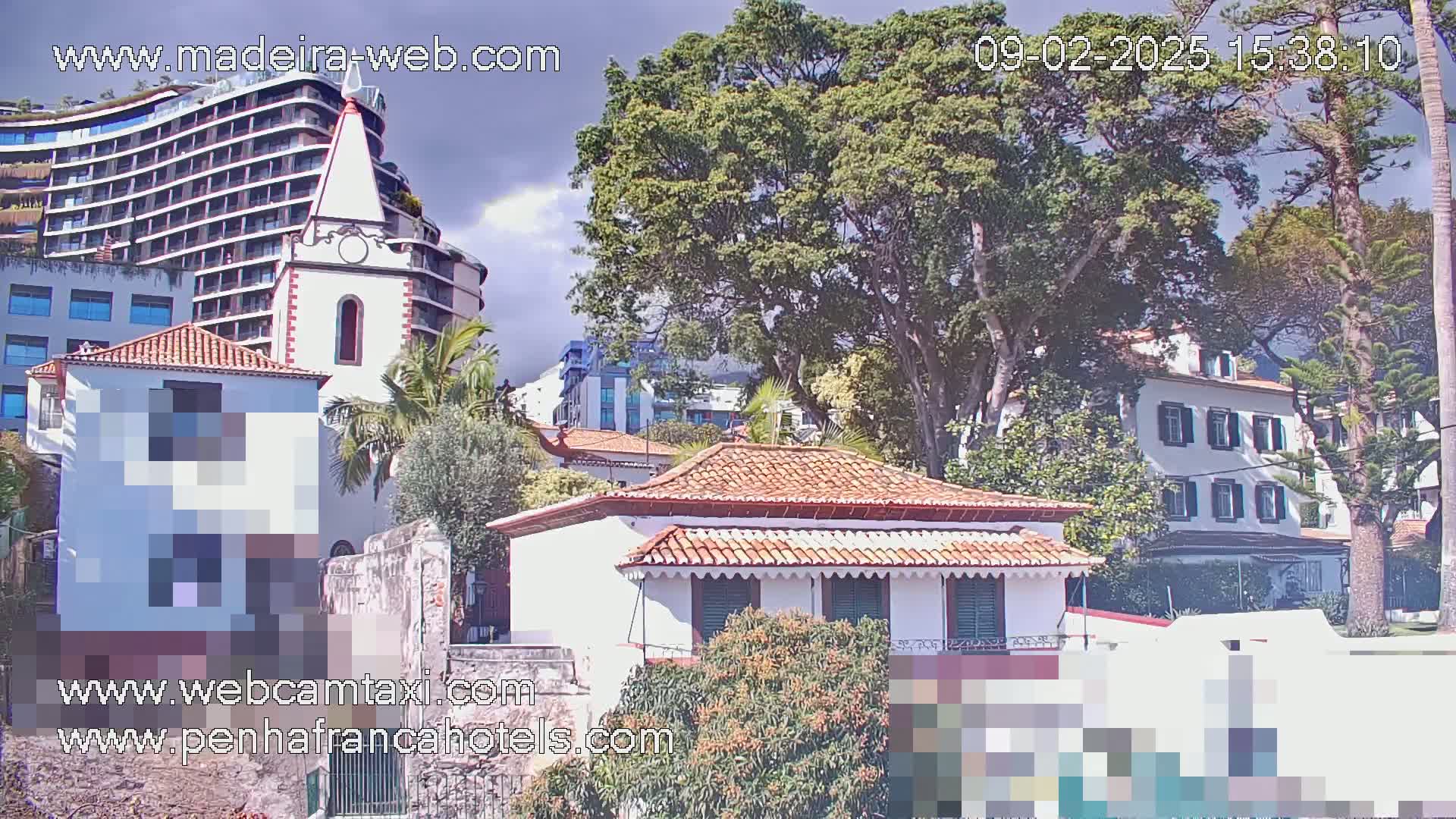 A partly cloudy day reveals several buildings, including a multi-story hotel, a white church, and smaller houses nestled among lush green trees.