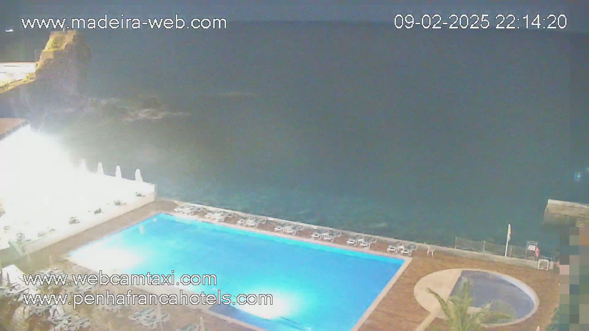 A nighttime view of an outdoor swimming pool next to a dark ocean, with waves splashing against the rocks below.