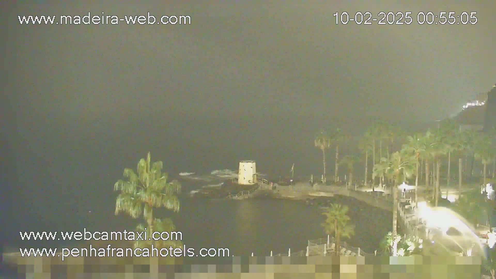A nighttime, hazy view of a rocky coast with palm trees, a small tower, and a calm body of water.