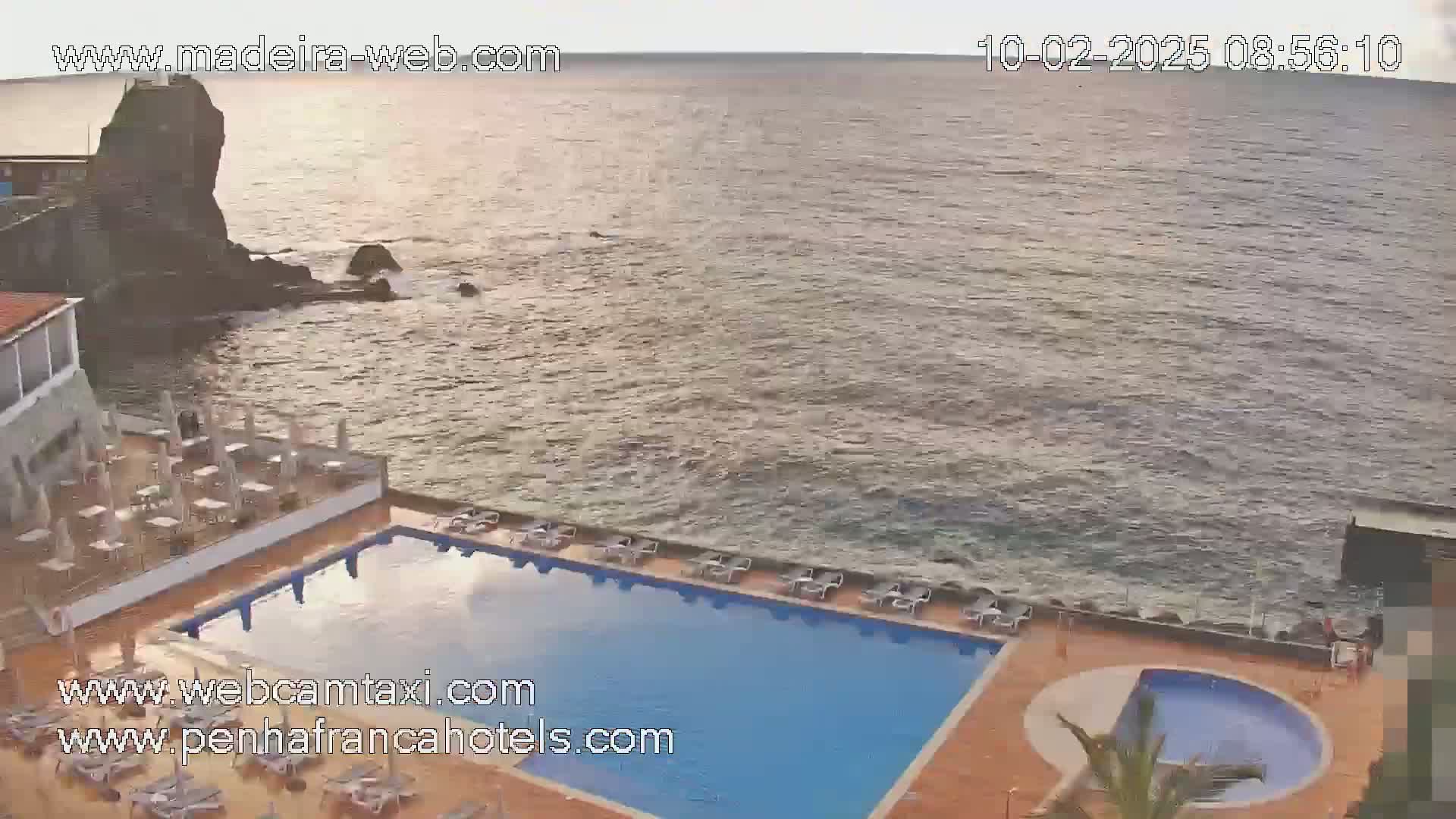 A rectangular swimming pool and a smaller round pool are situated next to a rocky ocean shore under a mostly cloudy sky.