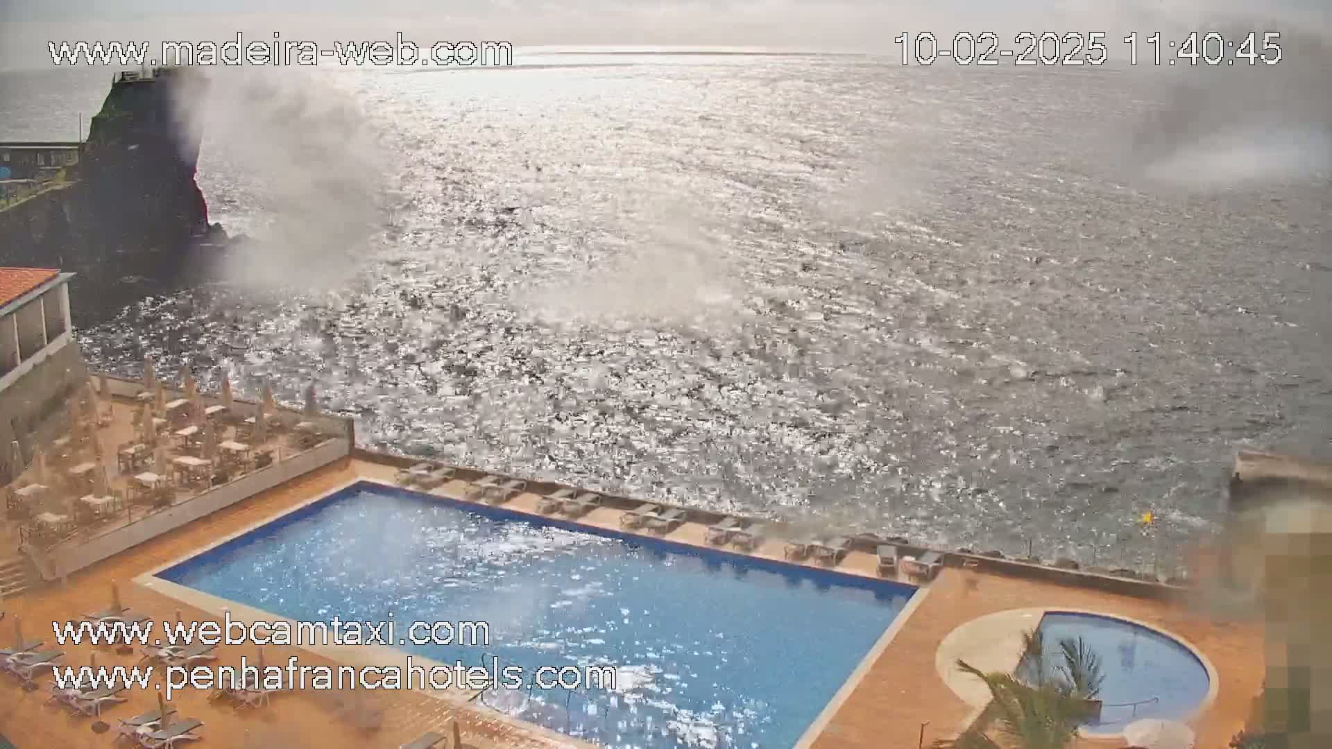 An outdoor swimming pool and a smaller circular pool are situated next to a rocky ocean shore on a windy day with choppy water.
