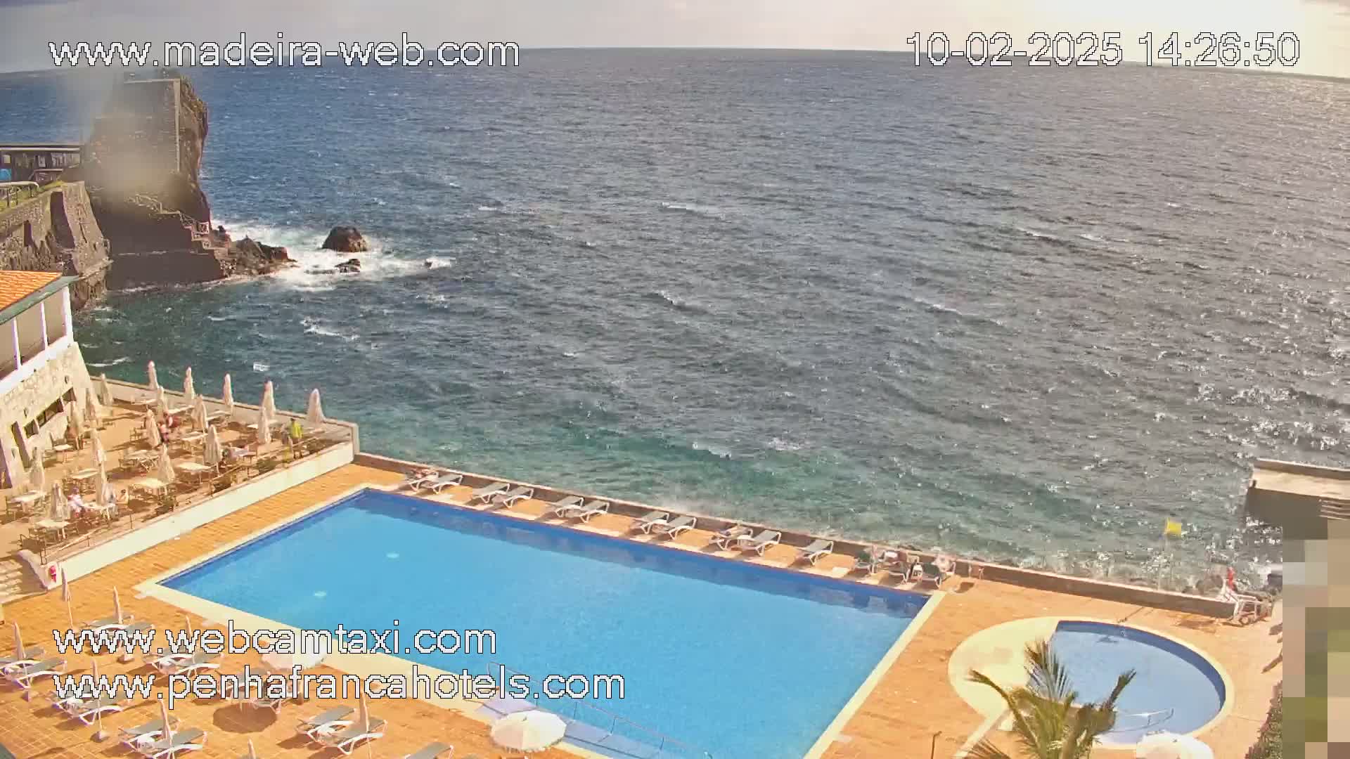 An outdoor swimming pool and lounge area are situated on a cliff overlooking a wavy ocean.