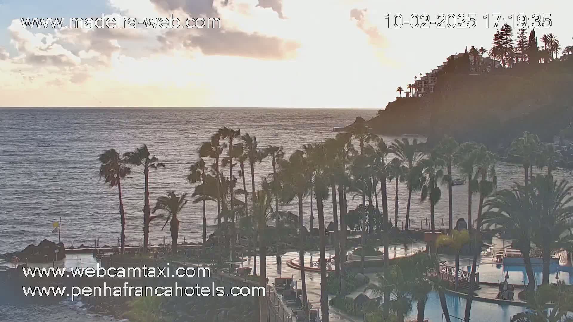 A partly cloudy sunset over a calm ocean, viewed from a resort with palm trees and swimming pools in the foreground and buildings on a cliff in the distance.