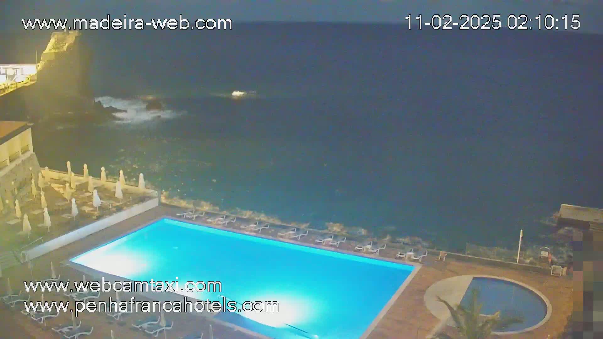 An illuminated swimming pool and smaller circular pool sit next to a dark ocean with small waves under a night sky.