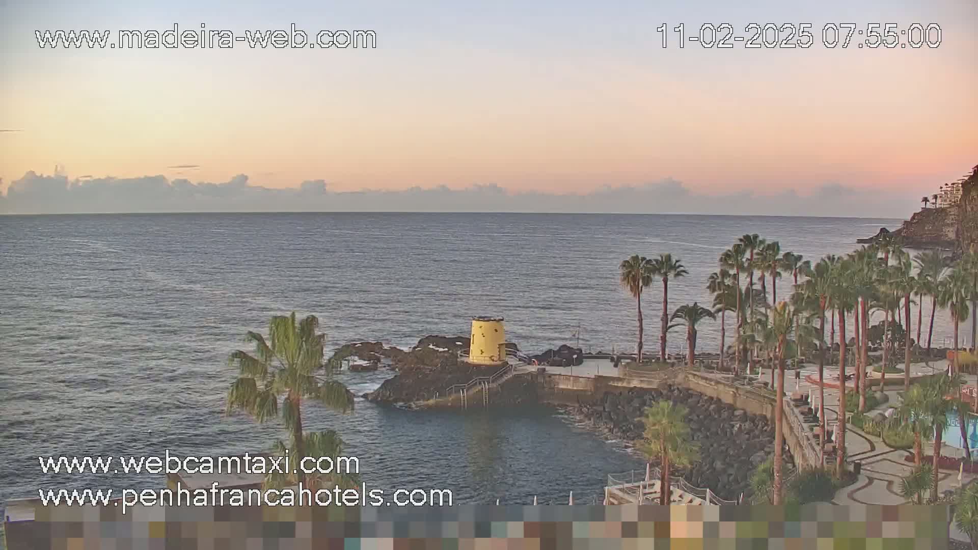A calm ocean at sunrise is viewed from a rocky coast with palm trees and a small yellow structure.