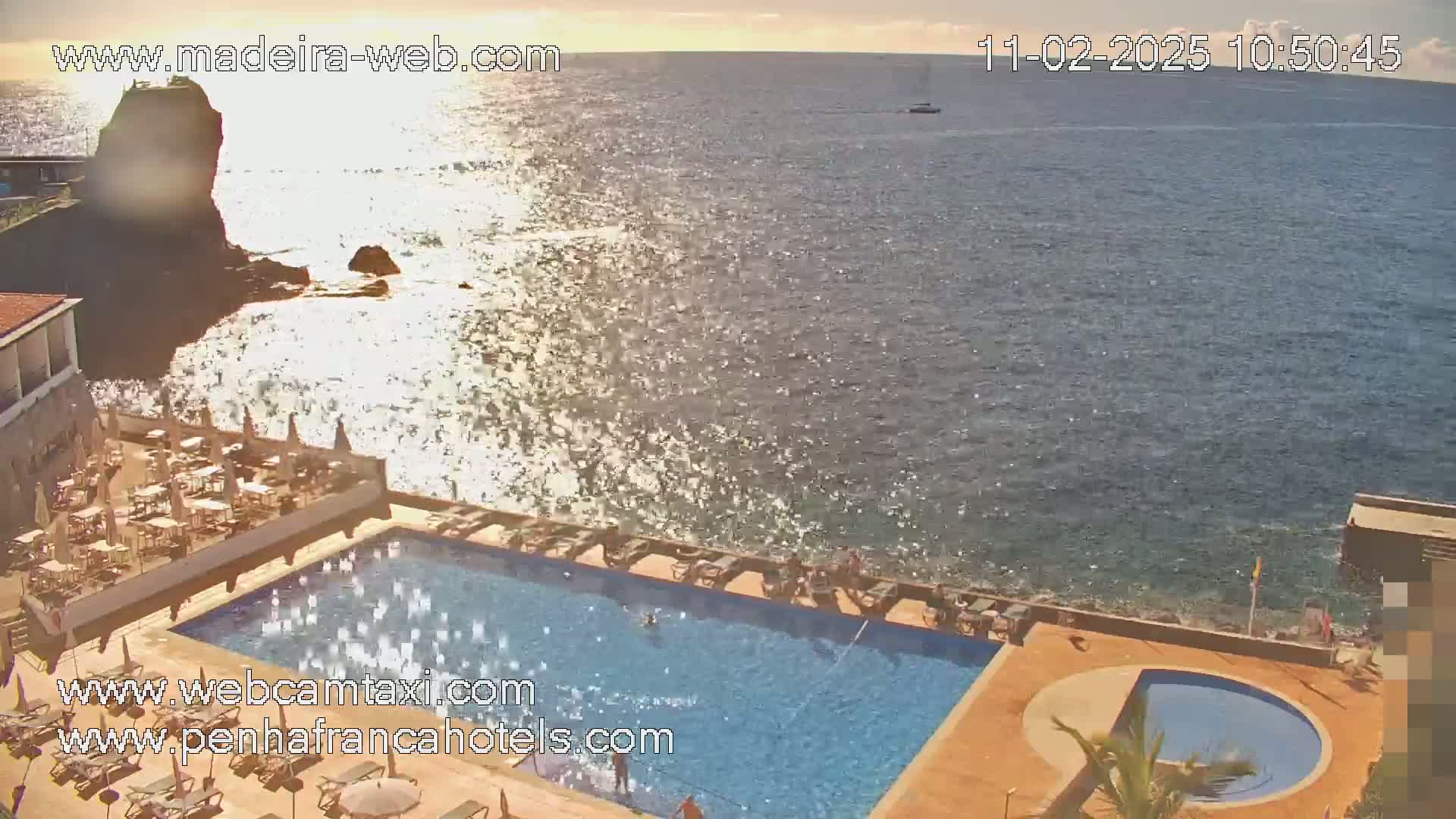 An aerial view shows a hotel pool and patio area next to a rocky coastline and a calm ocean under a partly sunny sky.