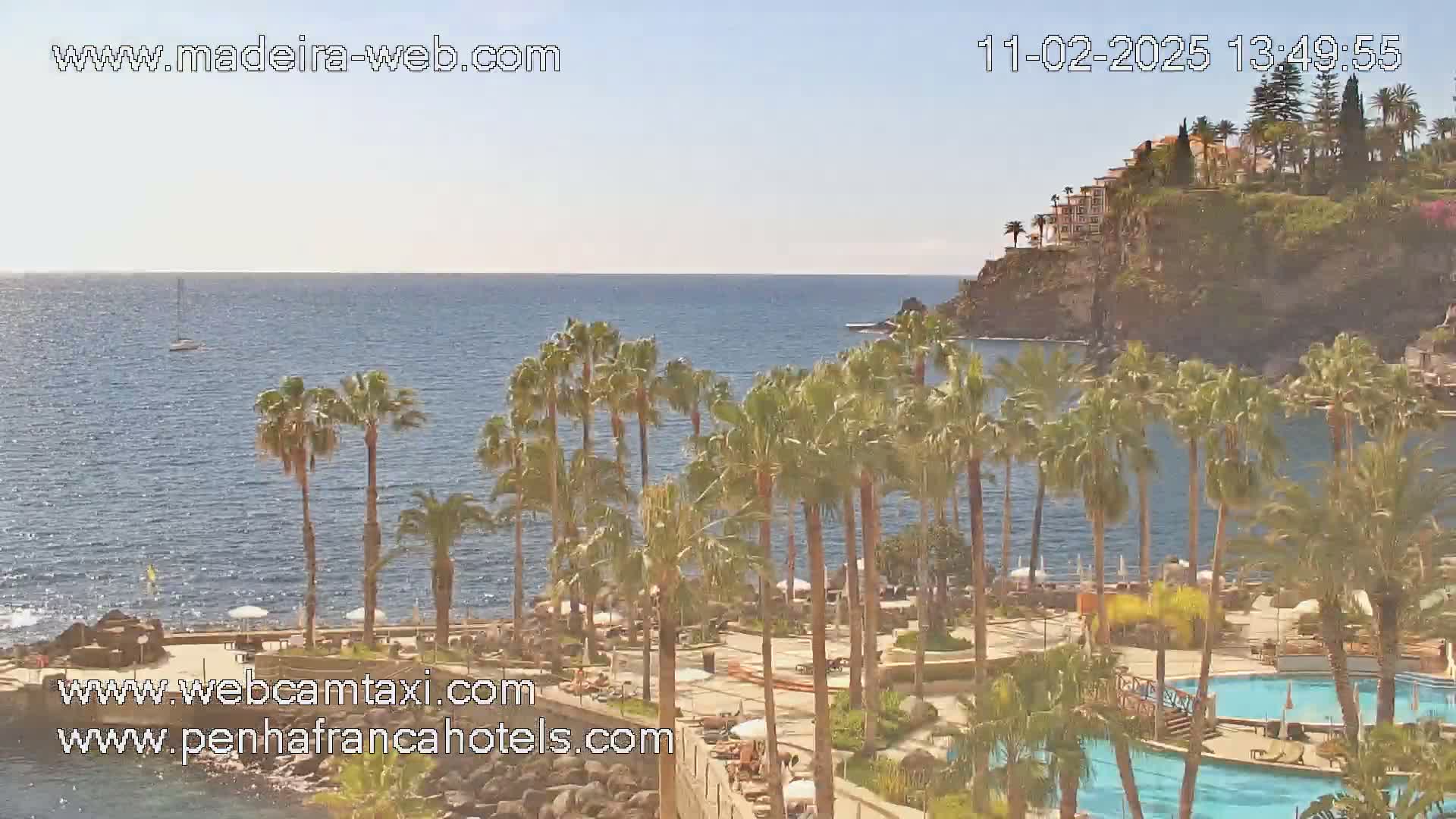 A resort with palm trees and swimming pools overlooks a calm ocean on a sunny day.
