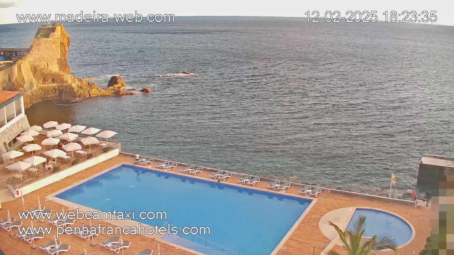 An outdoor swimming pool and patio area with umbrellas overlook a calm ocean next to a rocky cliff.