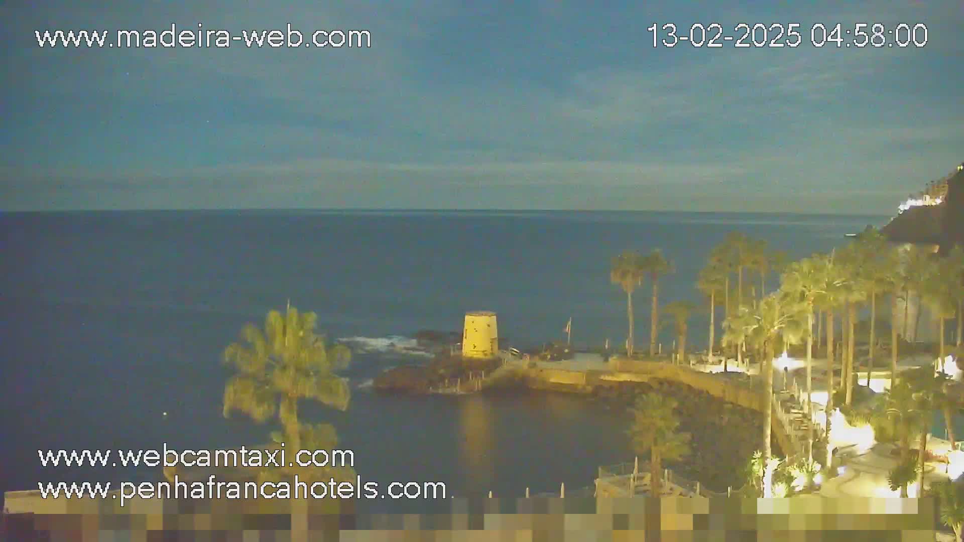 A nighttime coastal scene shows a calm ocean under a dusky sky, palm trees lining a walkway leading to a small, yellow tower on a rocky outcrop.