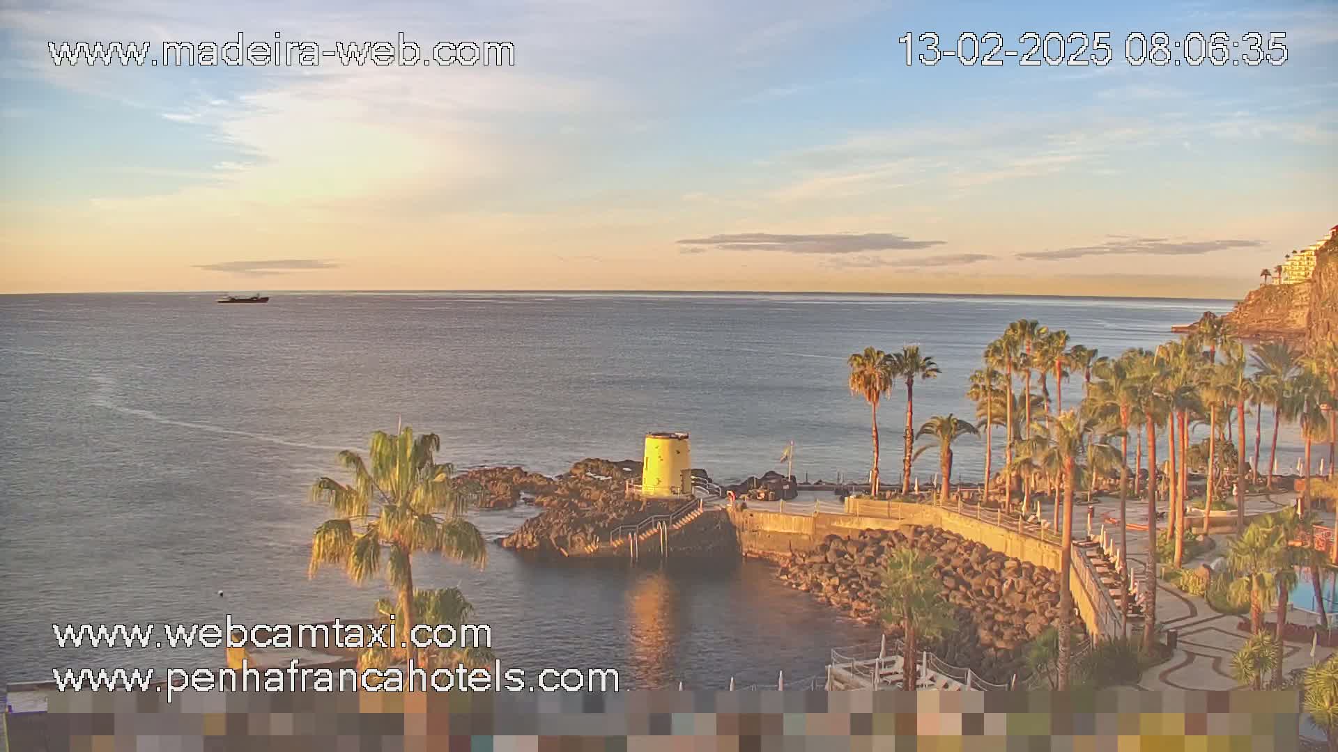A calm ocean with a distant ship is viewed from a rocky coast, where palm trees line a paved area near a small yellow structure under a partly cloudy, sunrise sky.