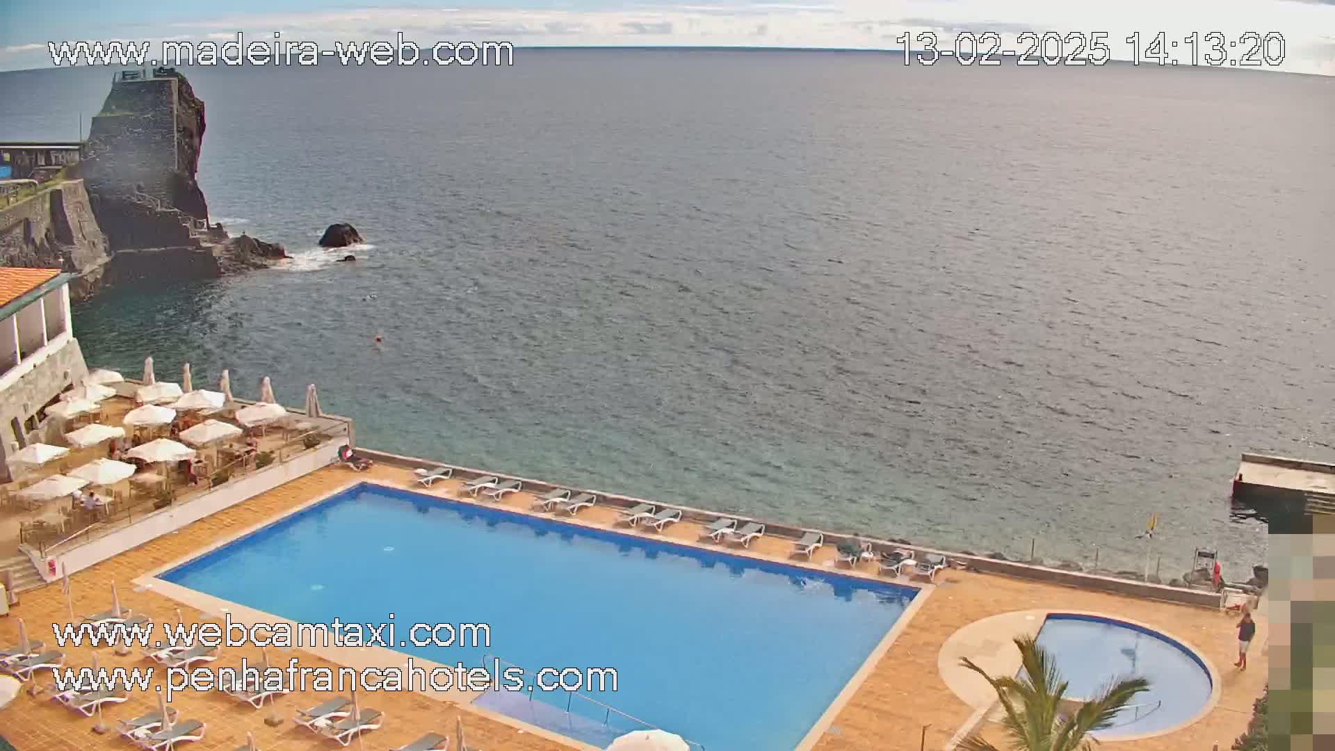 An outdoor swimming pool and smaller circular pool sit on a patio overlooking a calm ocean next to a rocky cliff on a partly cloudy day.