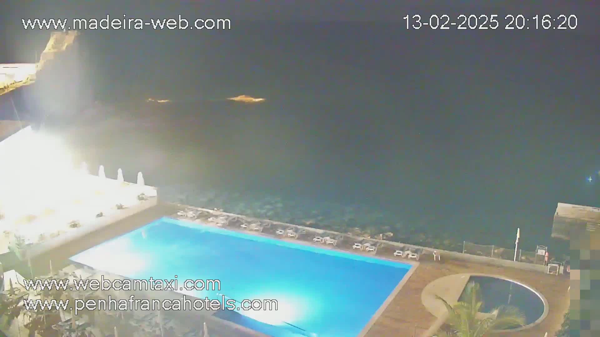An outdoor nighttime scene shows a large rectangular swimming pool illuminated next to a smaller circular pool, both situated on a deck overlooking a dark ocean.
