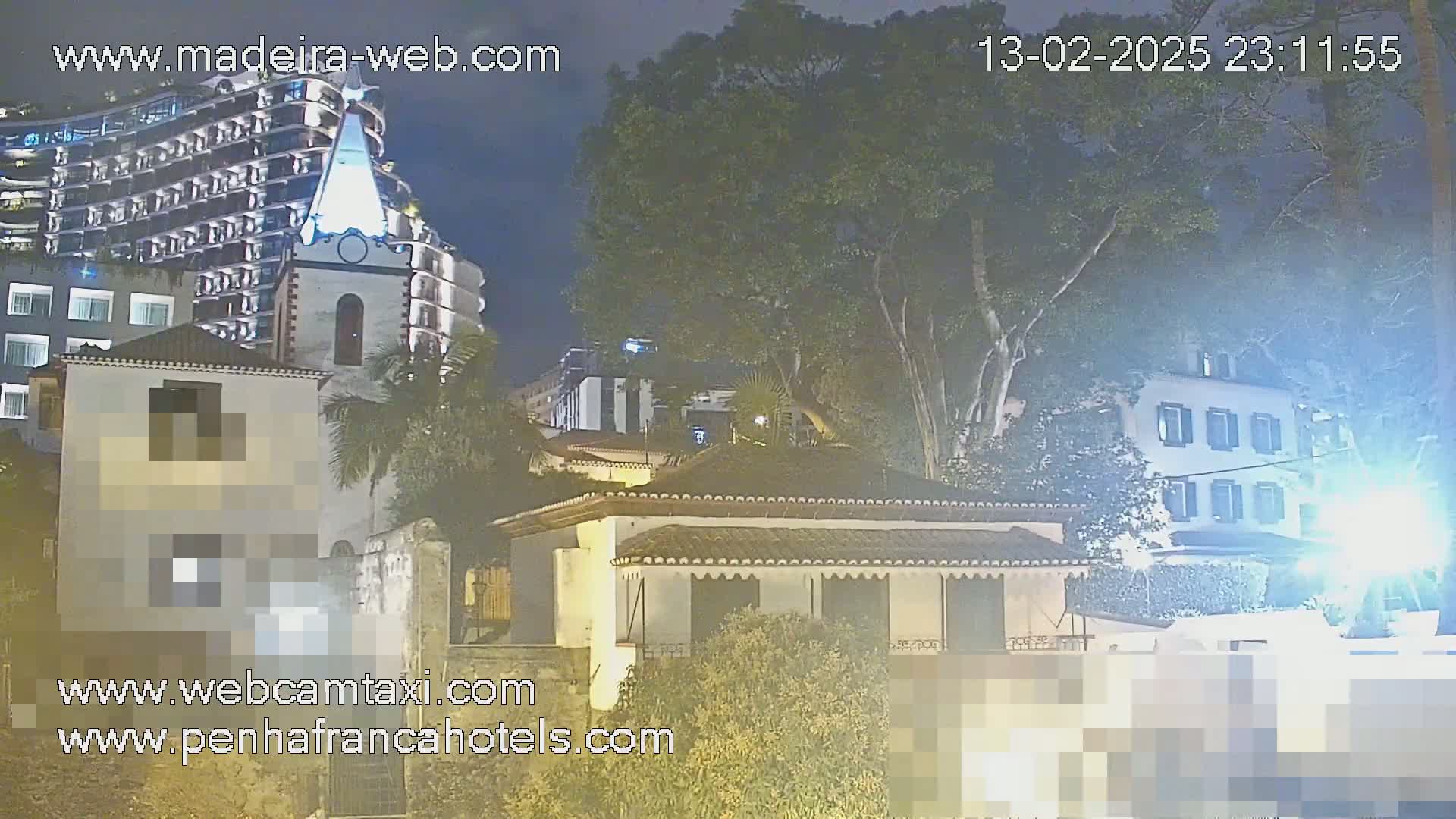 At night, under a dark sky, several buildings including a tall hotel and smaller houses are illuminated, with a large tree partially obscuring the view.