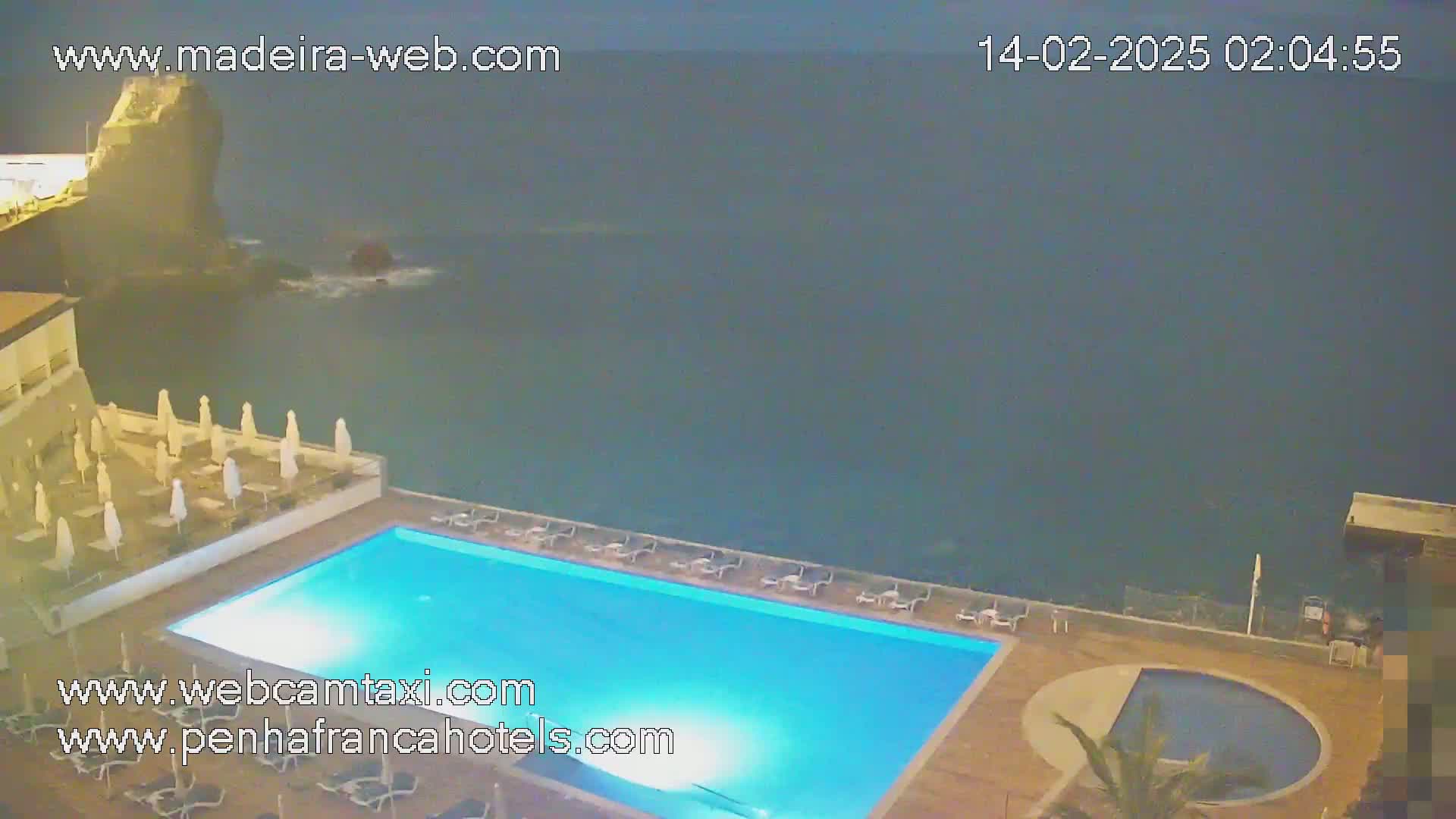 A nighttime view of a resort pool area with lounge chairs and umbrellas overlooking a dark ocean.
