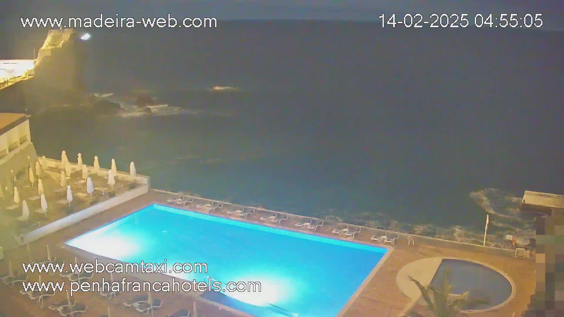 An illuminated swimming pool and surrounding patio area overlook a dark, wavy ocean at night.