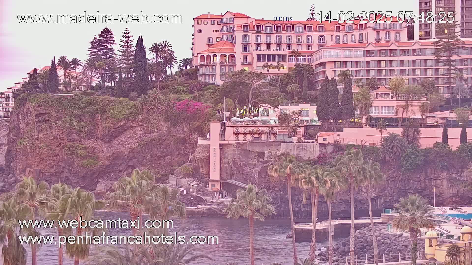 A large pink hotel complex sits atop a rocky cliff overlooking a calm body of water, surrounded by lush greenery and palm trees under a soft, dusky light.