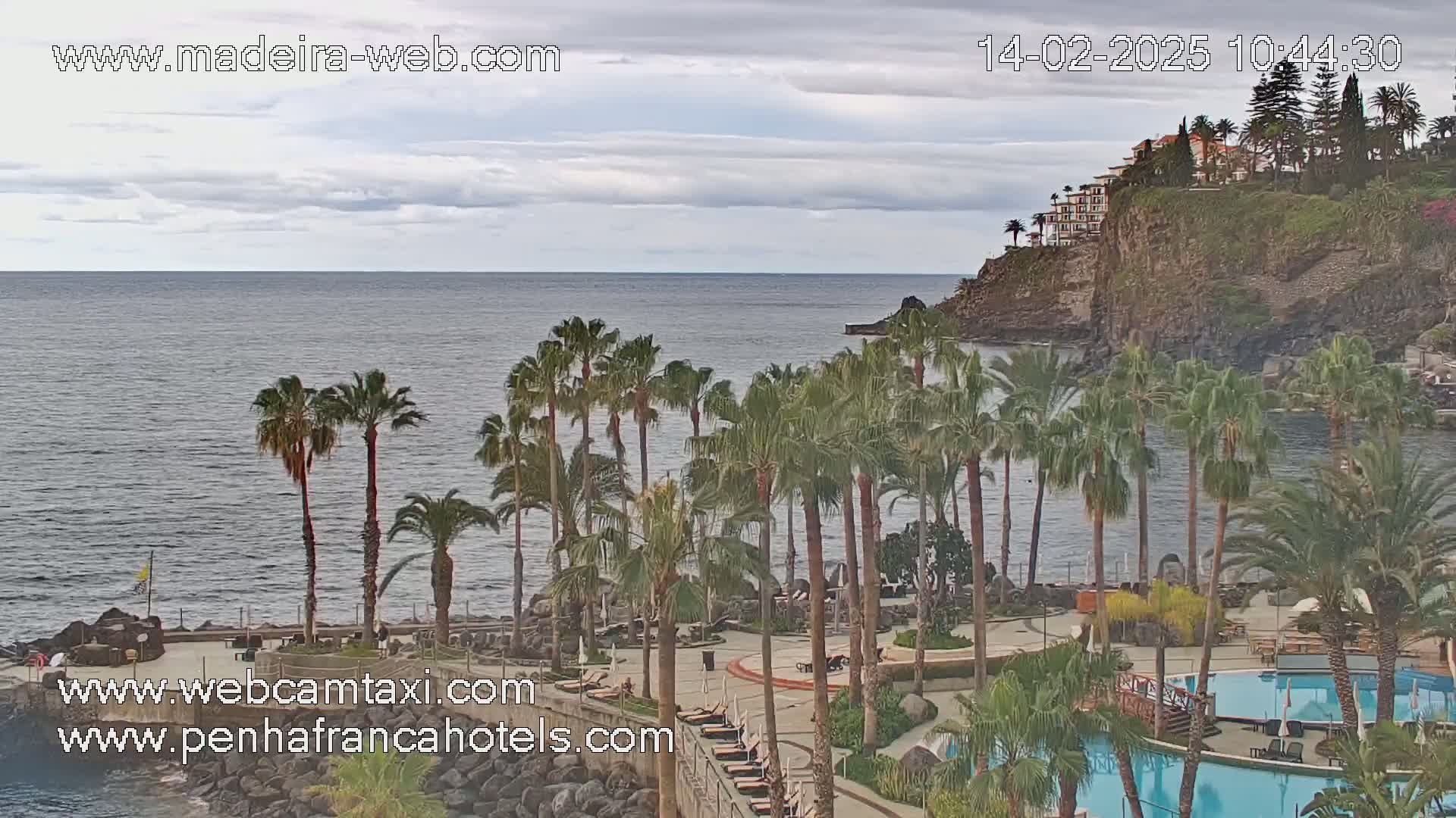 A resort with palm trees and swimming pools overlooks a calm ocean under a cloudy sky.