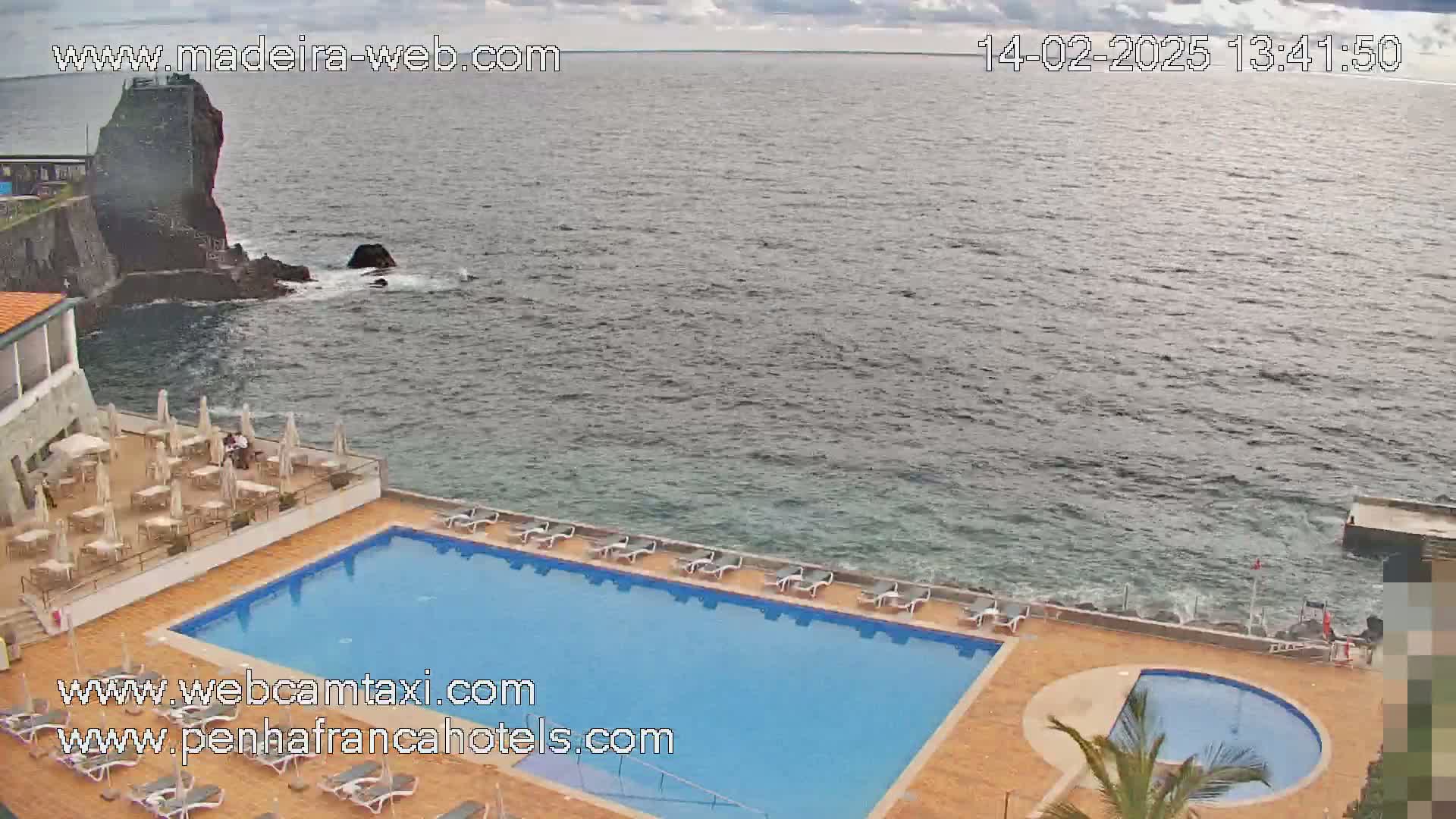 An outdoor pool with lounge chairs sits beside a dark-blue ocean on an overcast day, with a rocky outcrop visible in the distance.