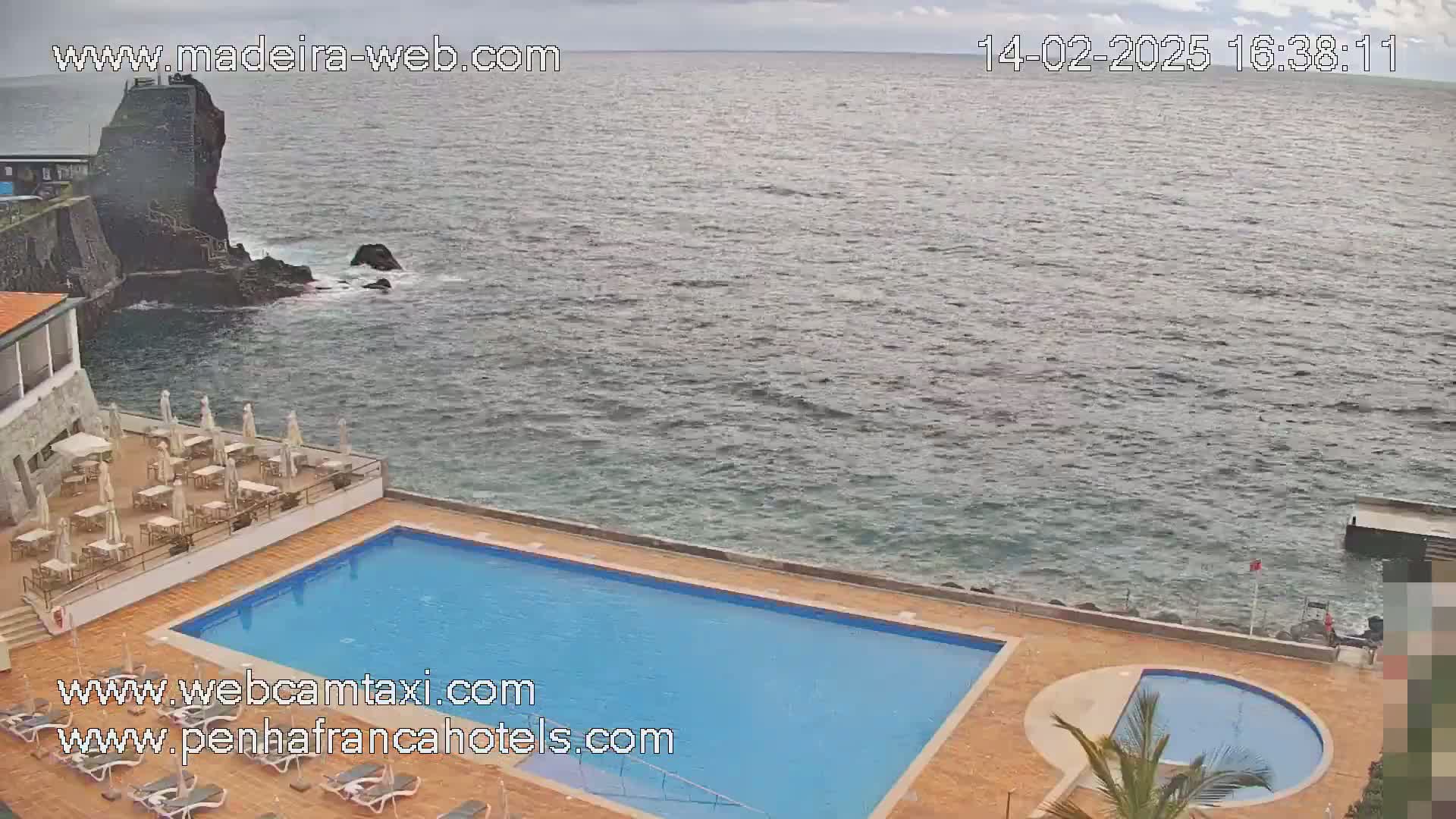 An outdoor pool and patio area overlooks a rocky coastline and choppy ocean under an overcast sky.