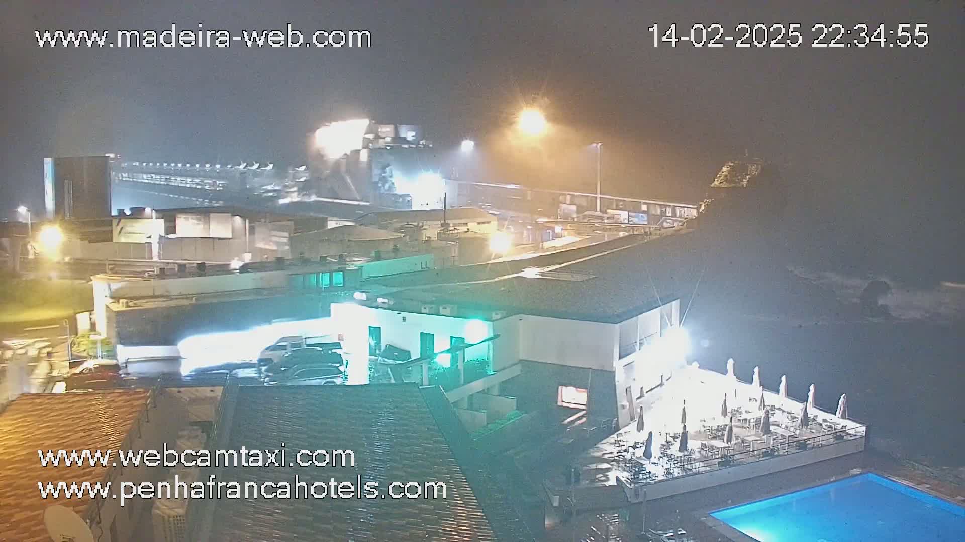 A nighttime view of a coastal town with rain or fog, showing buildings, vehicles, a pool, and ocean waves crashing against rocks.