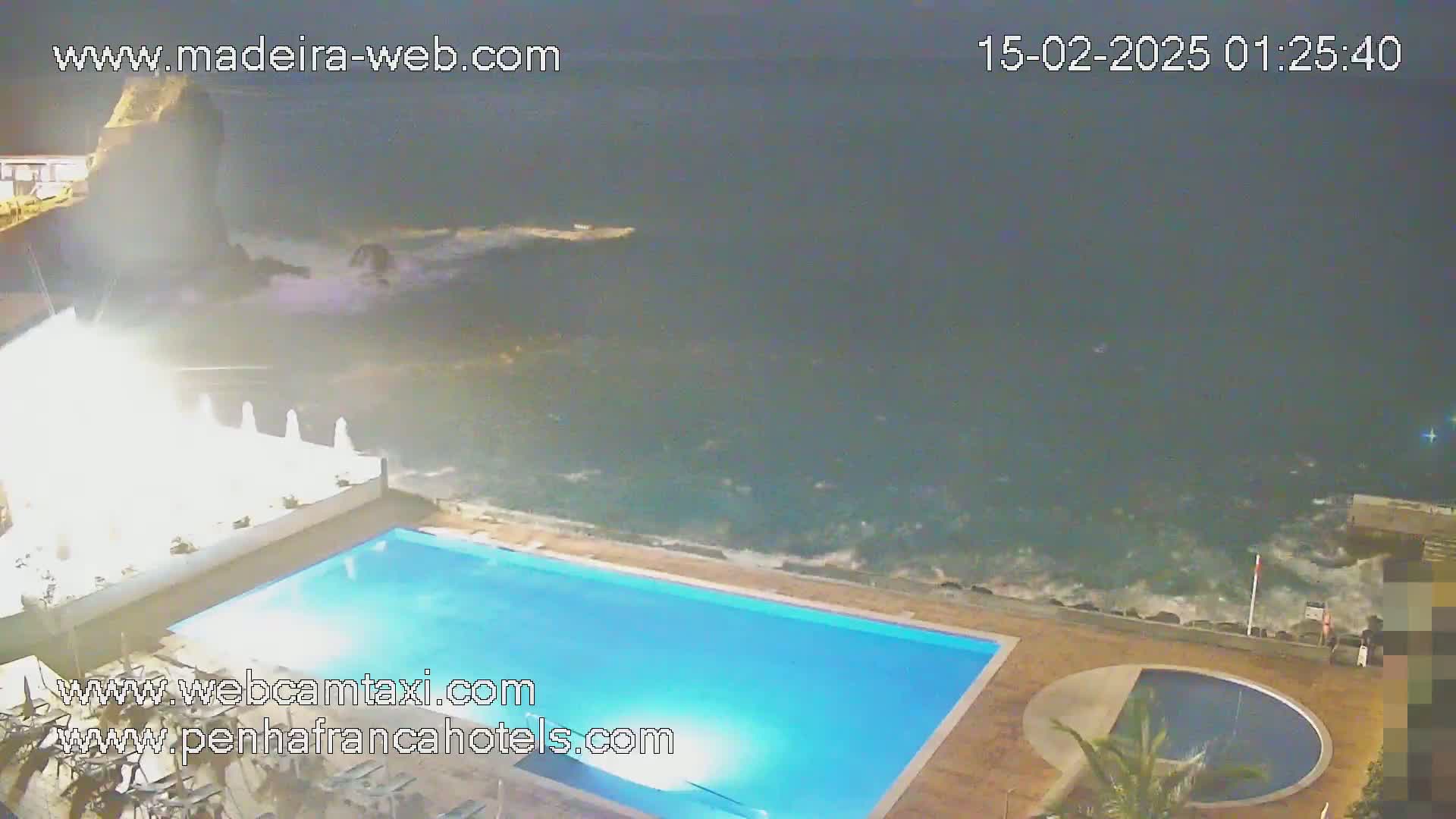 At night, a lit swimming pool sits next to a dark ocean with waves crashing against a rocky shore.